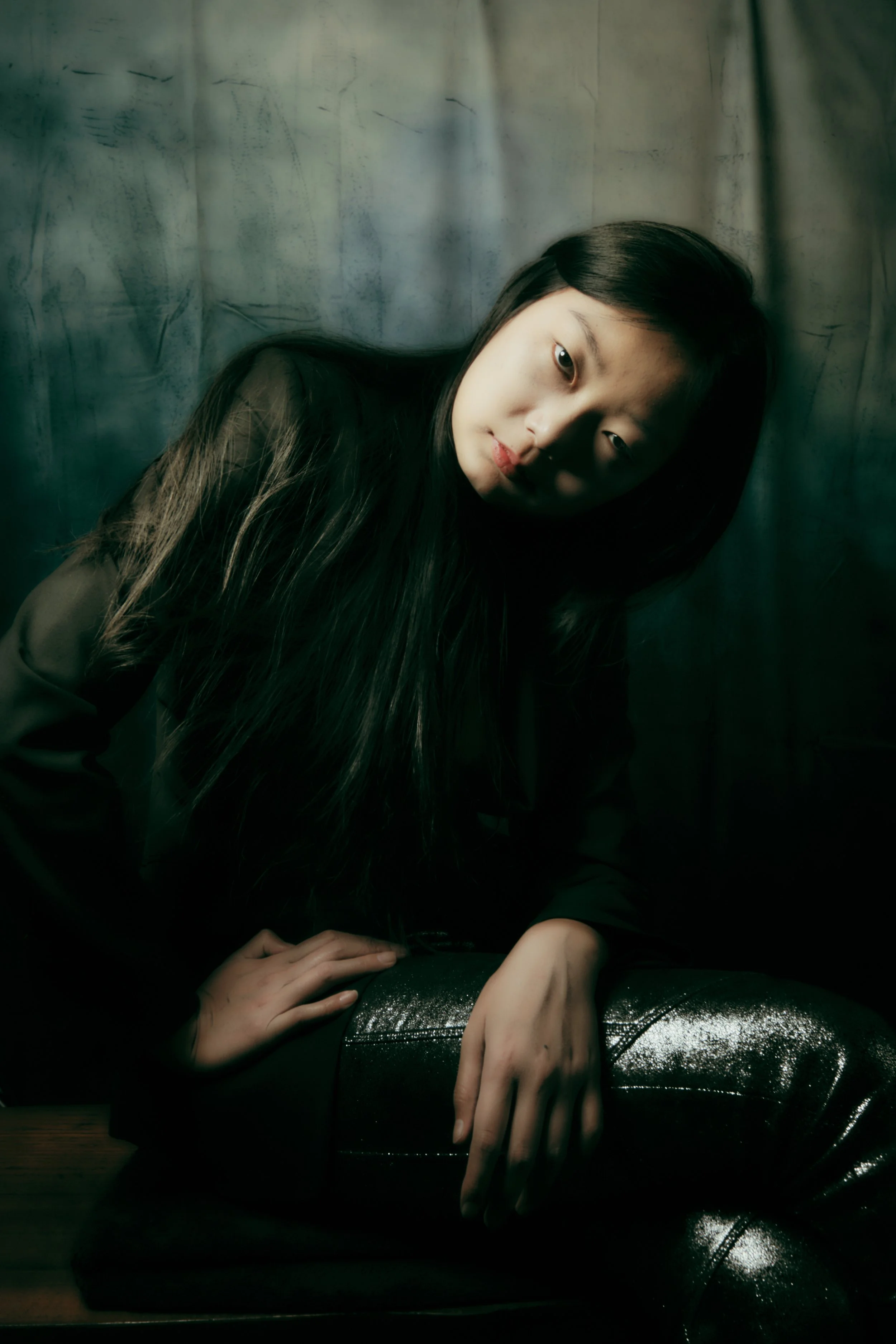 Portrait of a young woman with long dark hair, sitting on a wooden surface against a textured background, wearing a dark jacket and shiny black pants, looking at the camera with a contemplative expression. Portrait Photographer New York City