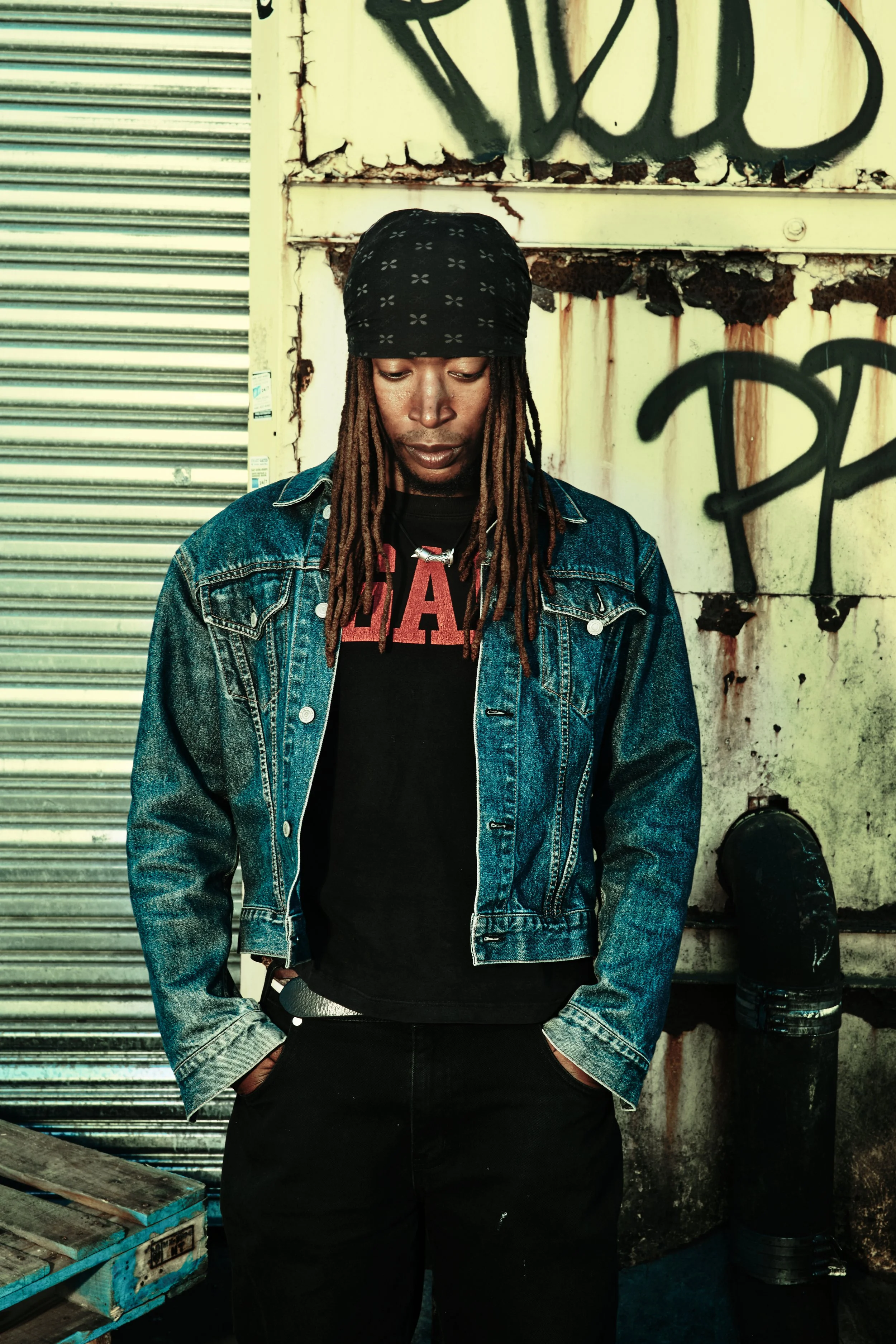 A man with dreadlocks wearing a bandana, denim jacket, and black t-shirt standing against a graffiti-covered, weathered wall in an urban setting. JSnaps Photography New York City Fashion lifestyle photography 