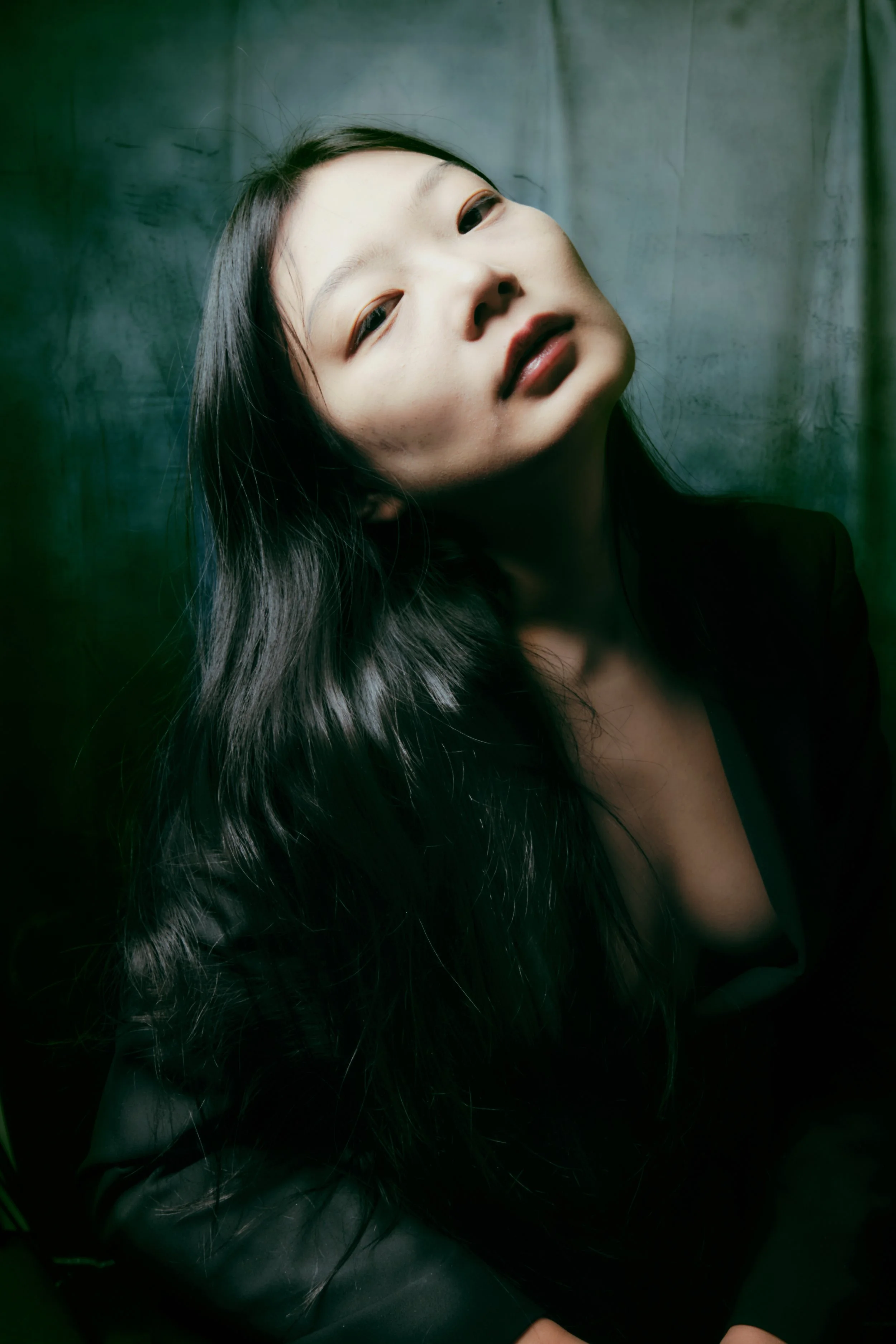 Portrait of a woman with long black hair, tilting her head with an intense expression, against a dark green background. Portrait Photographer New York City