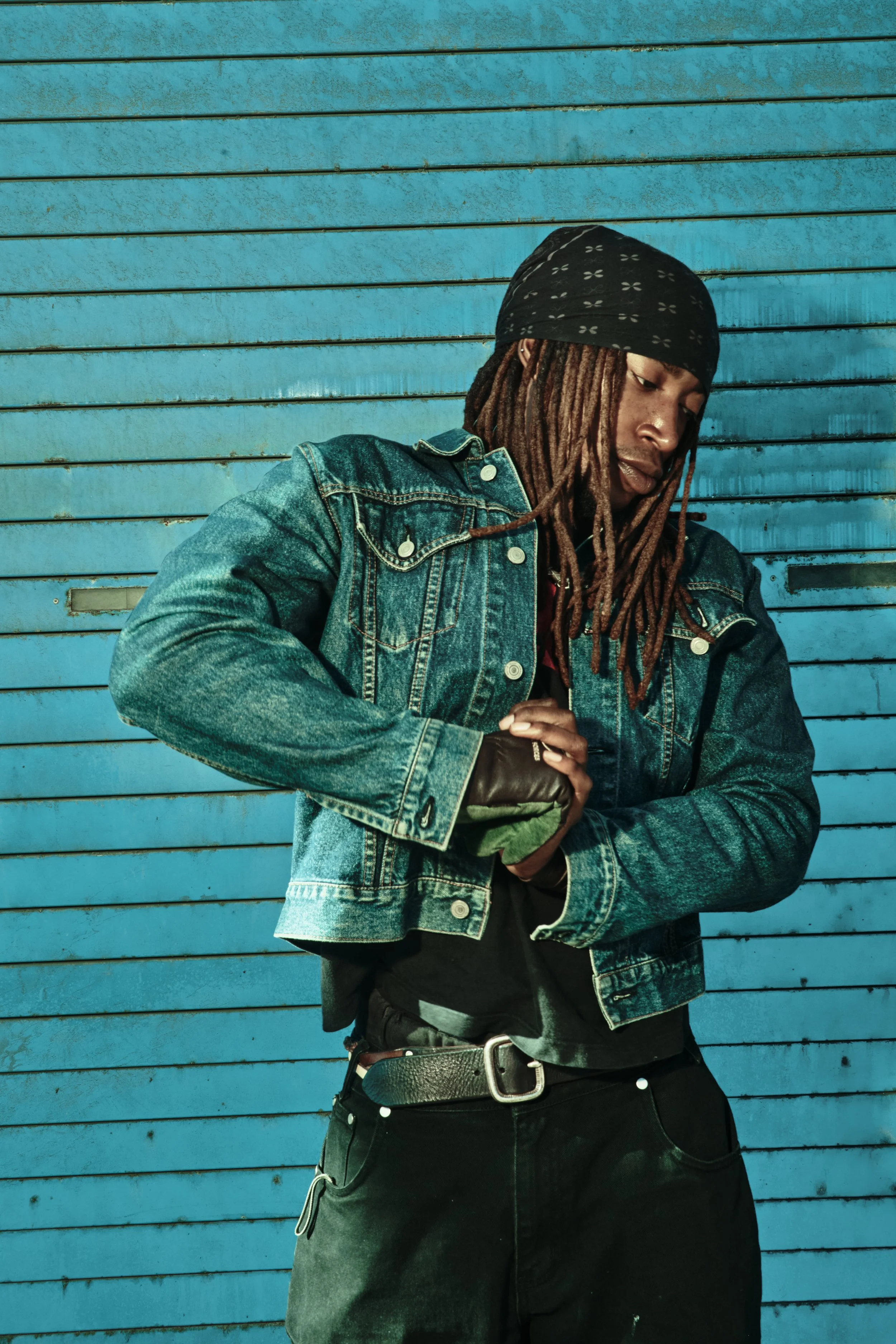 A young man with dreadlocks wearing a black bandana, denim jacket, black gloves, and black pants stands against a blue metal wall, looking down while holding what appears to be a green object. JSnaps Photography New York City Fashion lifestyle photog