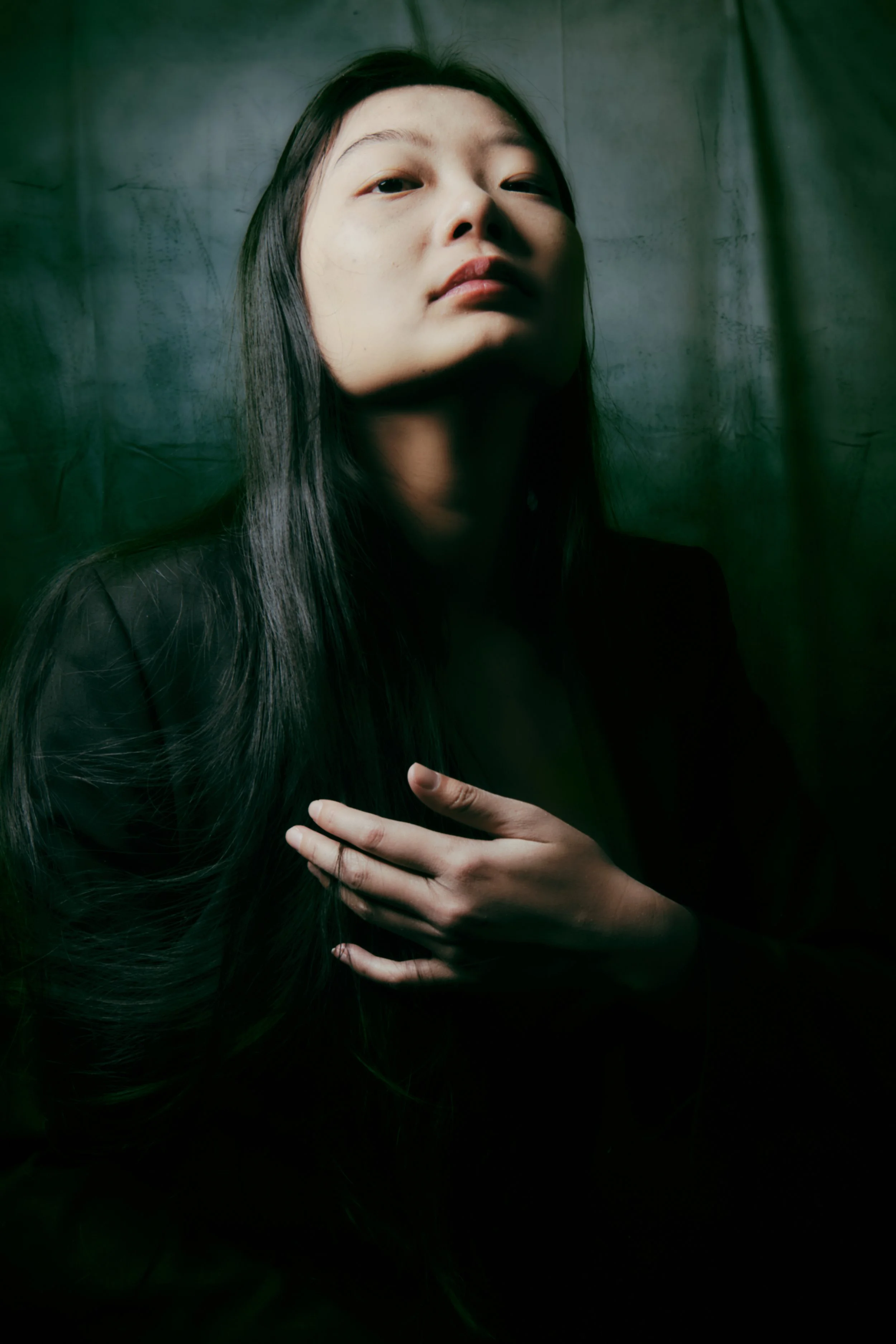 Portrait of a woman with long dark hair looking slightly upward, hand resting on chest, dark background with greenish tint. Portrait Photographer New York City