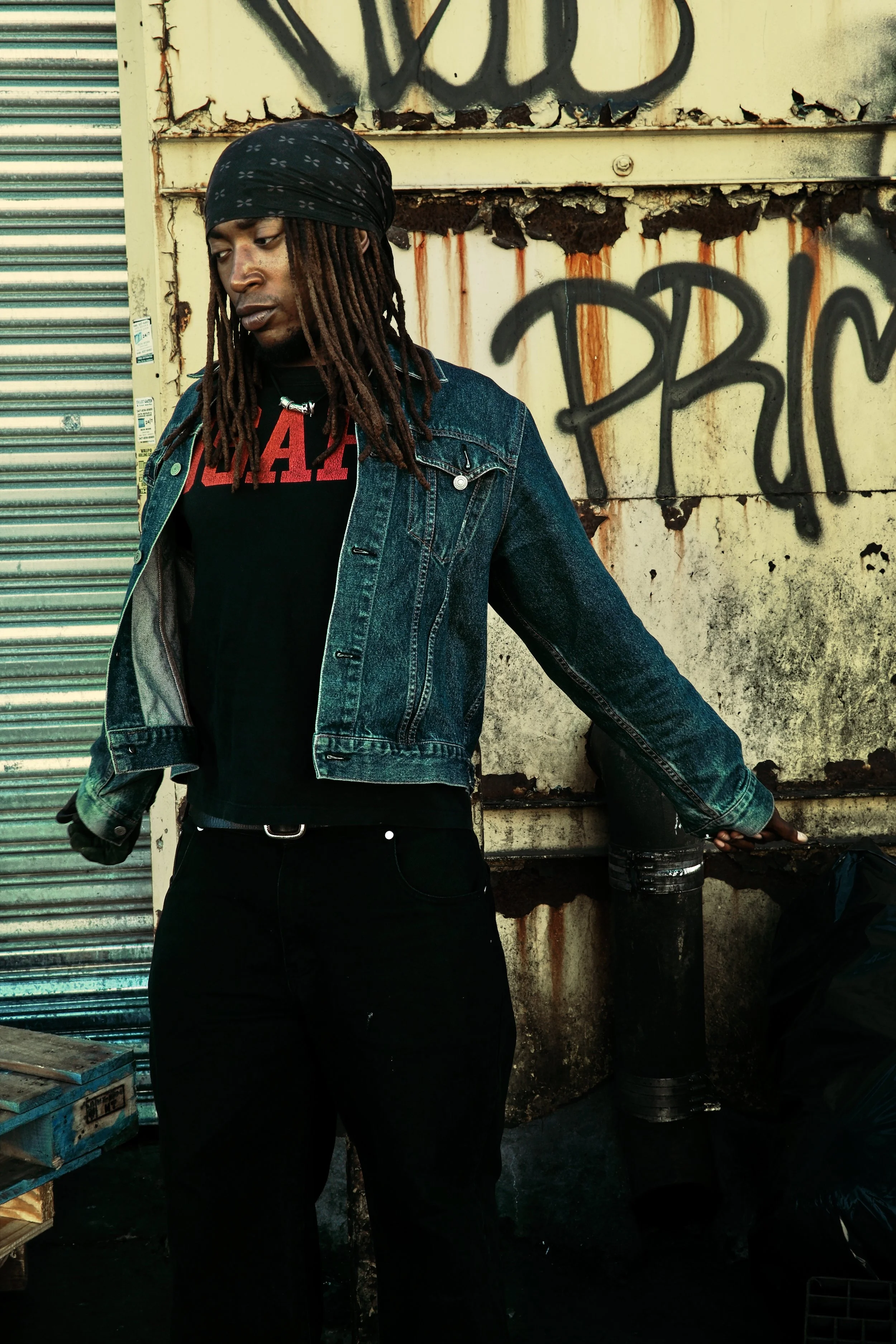 A young man with dreadlocks and a black bandana standing outdoors against a graffiti-covered, rusted metal wall, wearing a denim jacket over a black shirt, with black pants and gloves. JSnaps Photography New York City Fashion lifestyle photography 