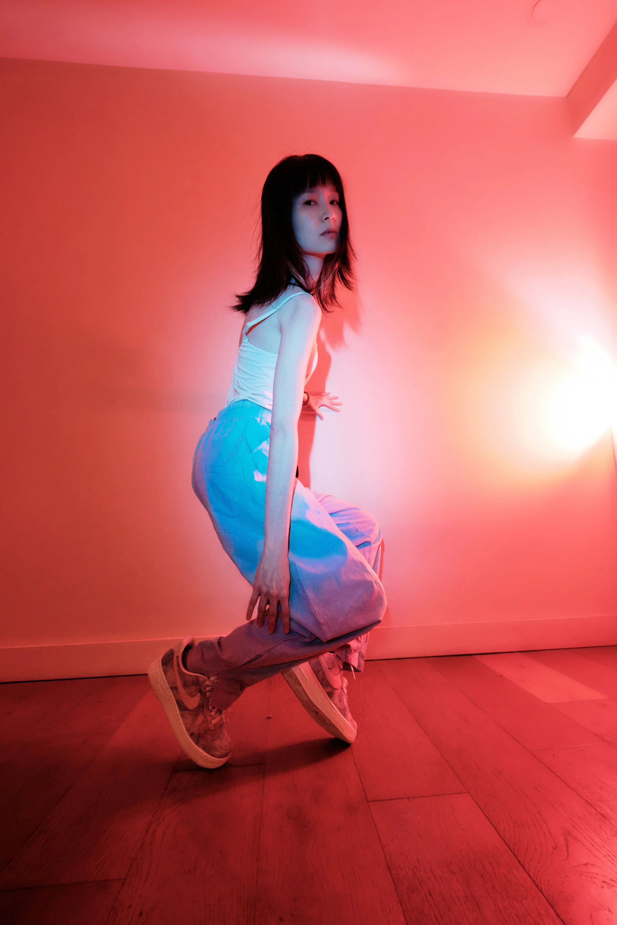 A young woman with black hair and bangs posing in a room with pink and red lighting. She is wearing a white sleeveless top, loose light blue pants, and sneakers. The floor is wooden, and the wall behind her is plain. She is crouching, with one hand o