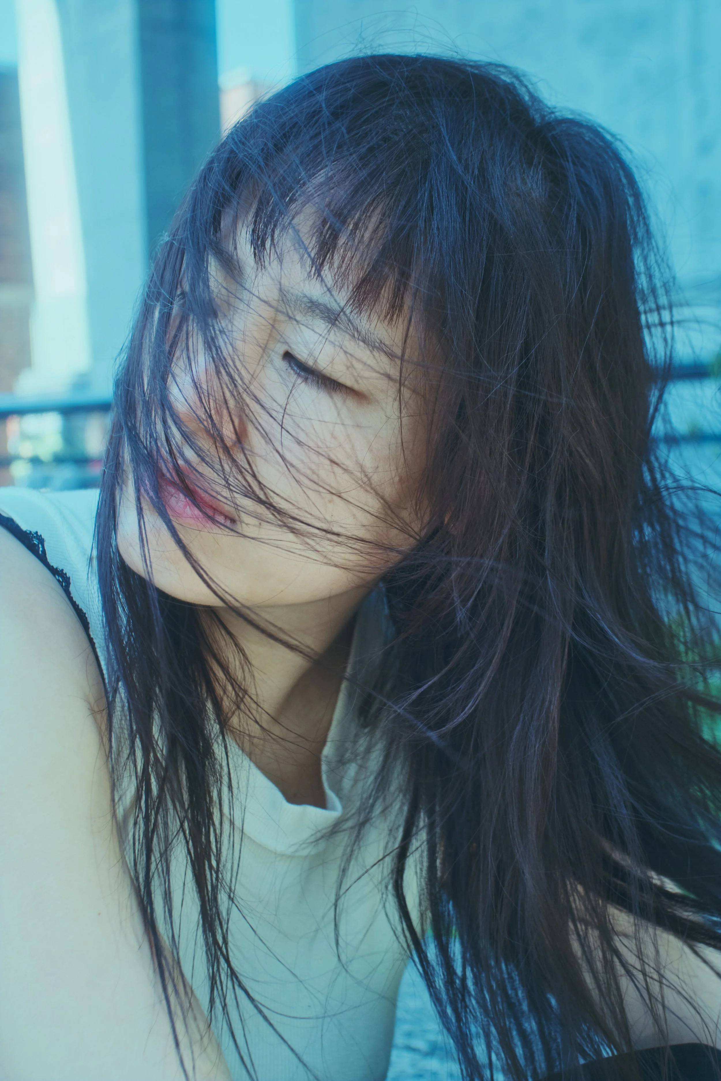 Close-up of a woman with long, dark hair blowing across her face, eyes closed, in front of a window with blue tones. JSnaps Photography New York City Fashion lifestyle photography 