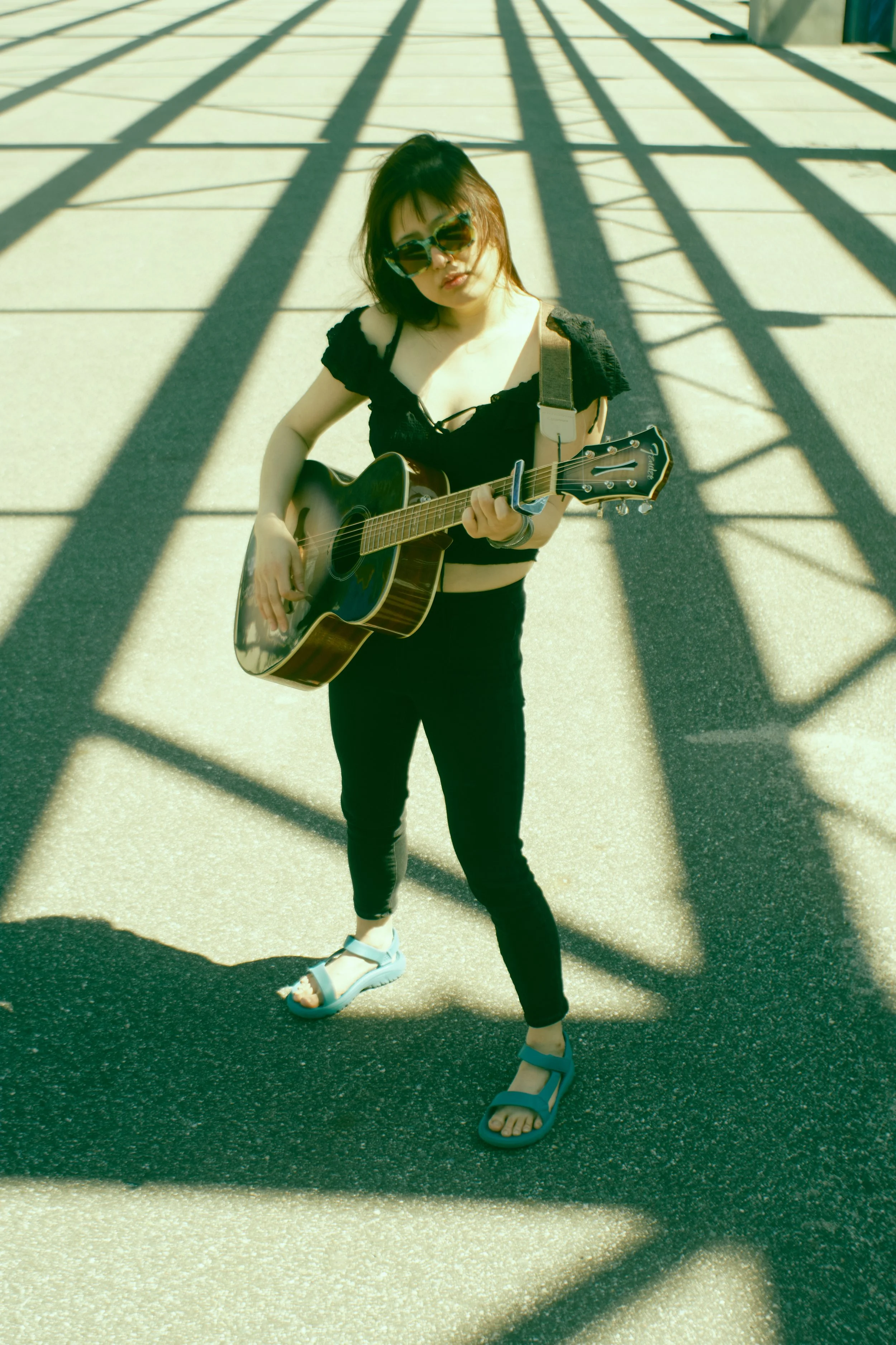 Young woman with dark hair and sunglasses playing an acoustic guitar outdoors on a sidewalk, with shadows of a metal structure overhead. JSnaps Photography New York City Fashion lifestyle photography 