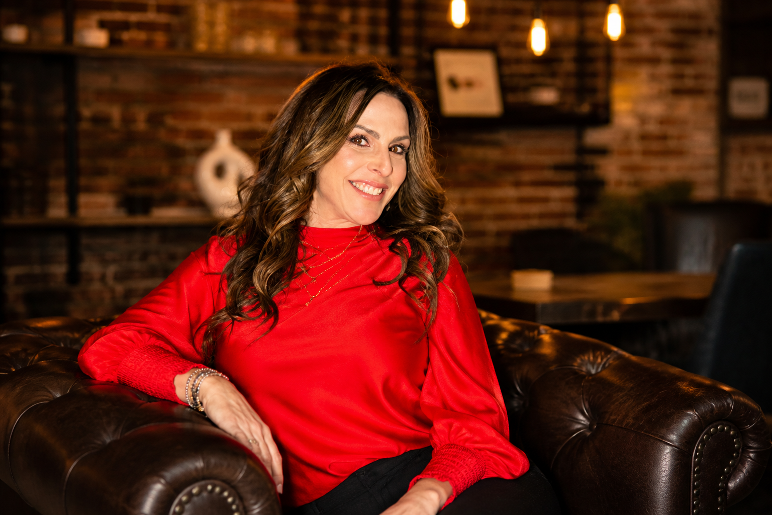 A woman with long wavy brown hair sitting on a dark brown leather Chesterfield sofa, wearing a red satin blouse, smiling with a brick wall background, decorated with wall shelves and hanging Edison-style light bulbs.