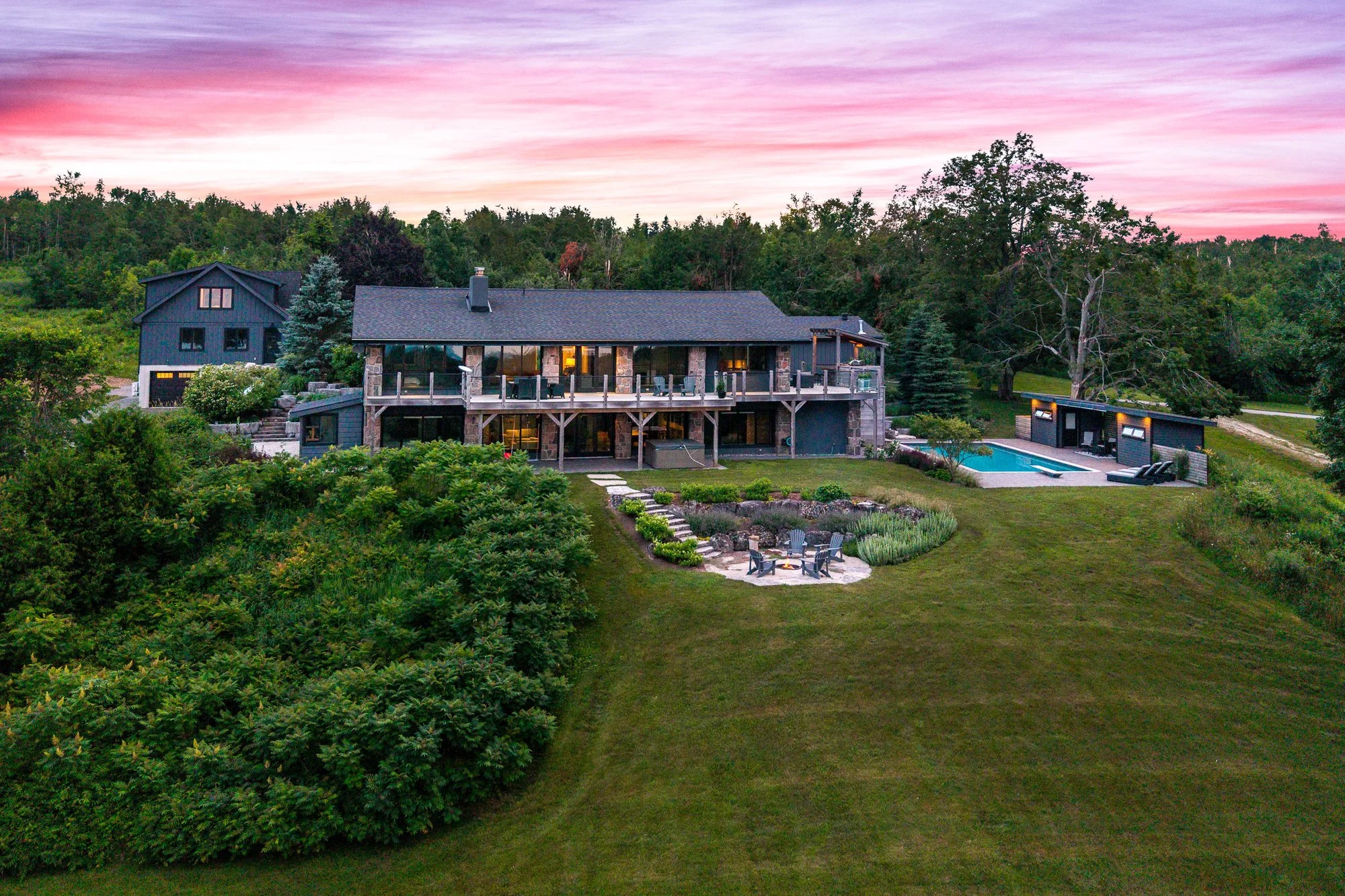A modern house with a large deck, a swimming pool, and a fire pit area in a lush green landscape during sunset with pink and purple clouds.