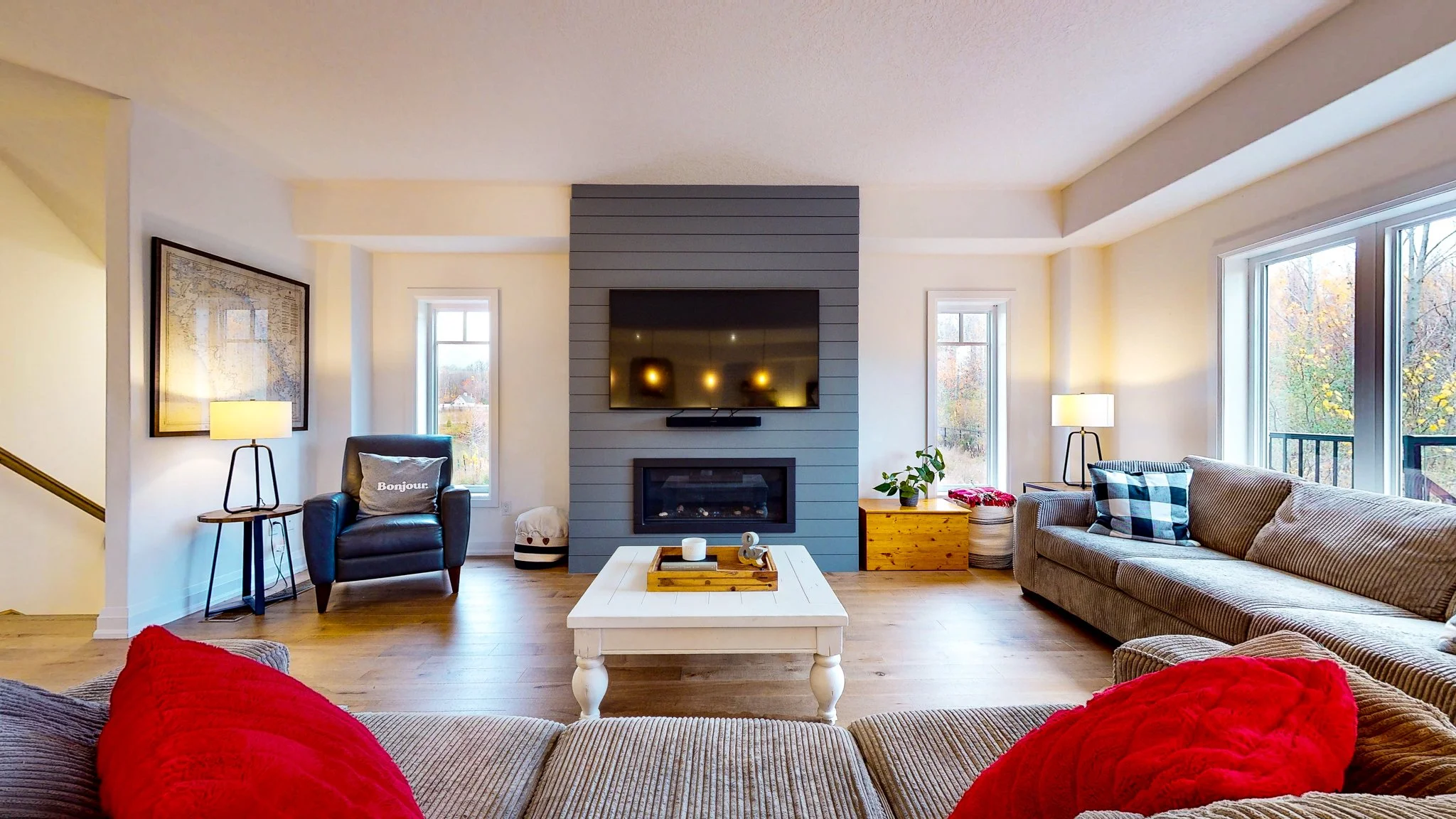 Living room with beige sofa, red pillows, white coffee table, fireplace, wall-mounted TV, armchair with pillow labeled "Bonjour", side tables with lamps, large windows with outdoor view of autumn trees.
