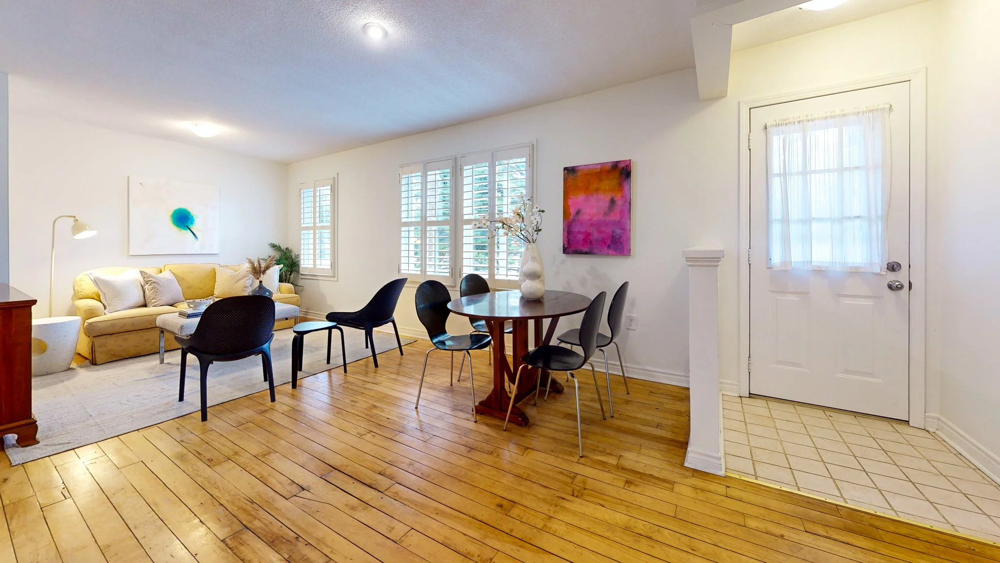 Living room and dining area with hardwood floors, white walls, yellow sofa, black chairs, and colorful abstract artwork.