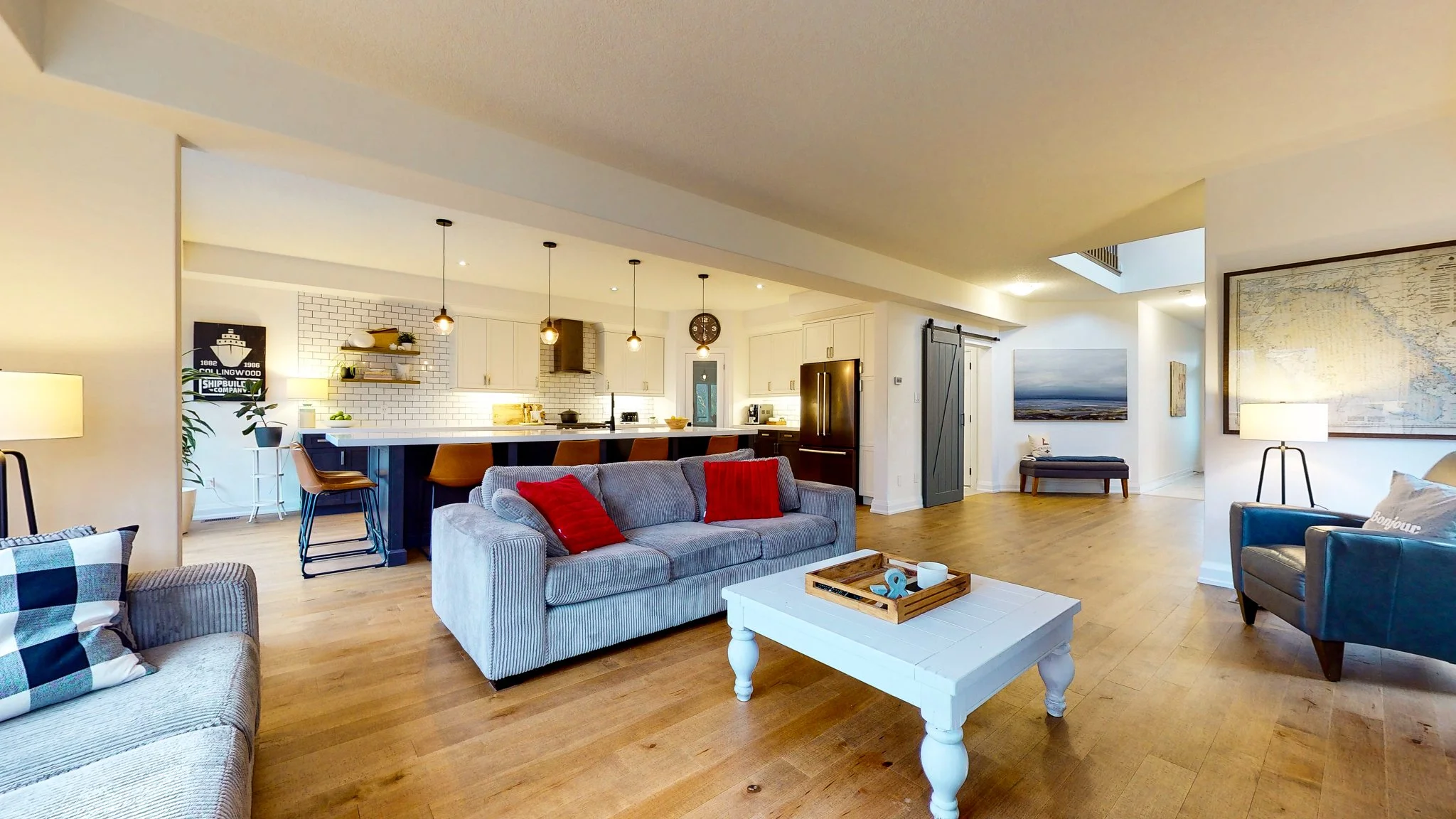 Open-concept living room and kitchen with hardwood floors, gray sofa with red pillows, blue armchair, white coffee table, and kitchen with white cabinets, black countertops, and pendant lighting.