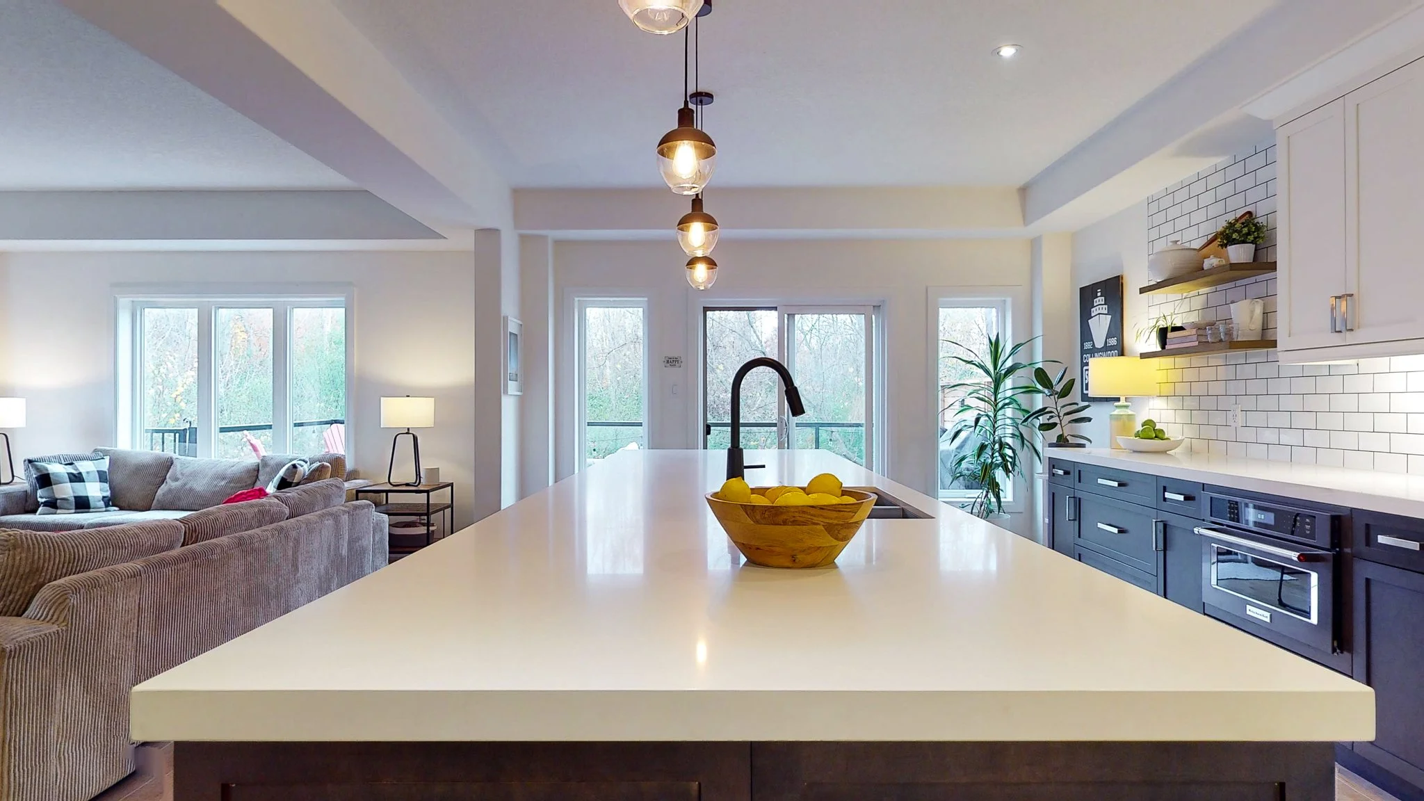 Modern open-concept kitchen with a white island, black fixtures, and blue cabinets, adjacent to a living room with beige sofas and large windows.