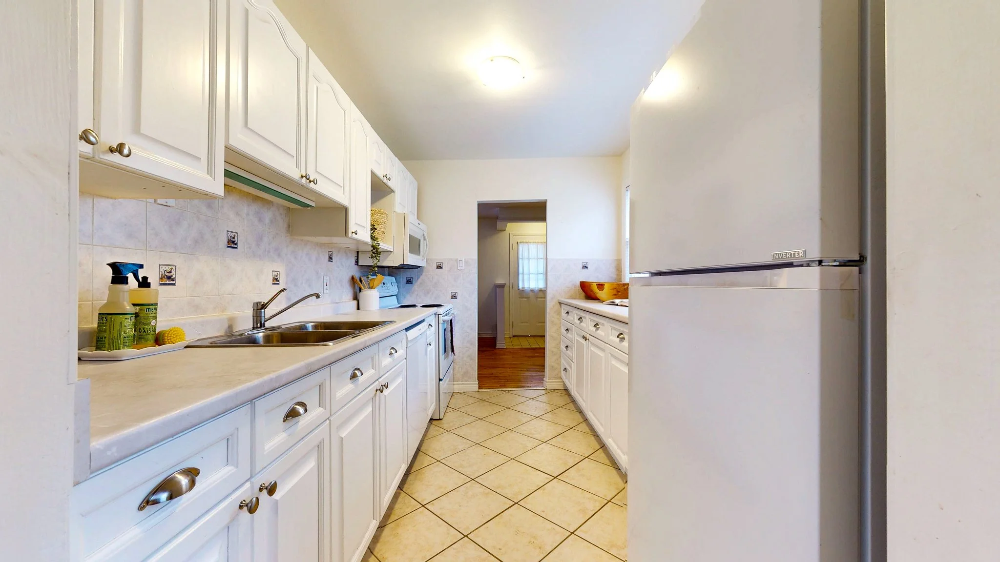 Bright kitchen with white cabinets, beige tiled floor, white refrigerator, stainless steel sink, and a doorway leading to a room with hardwood flooring and a window.