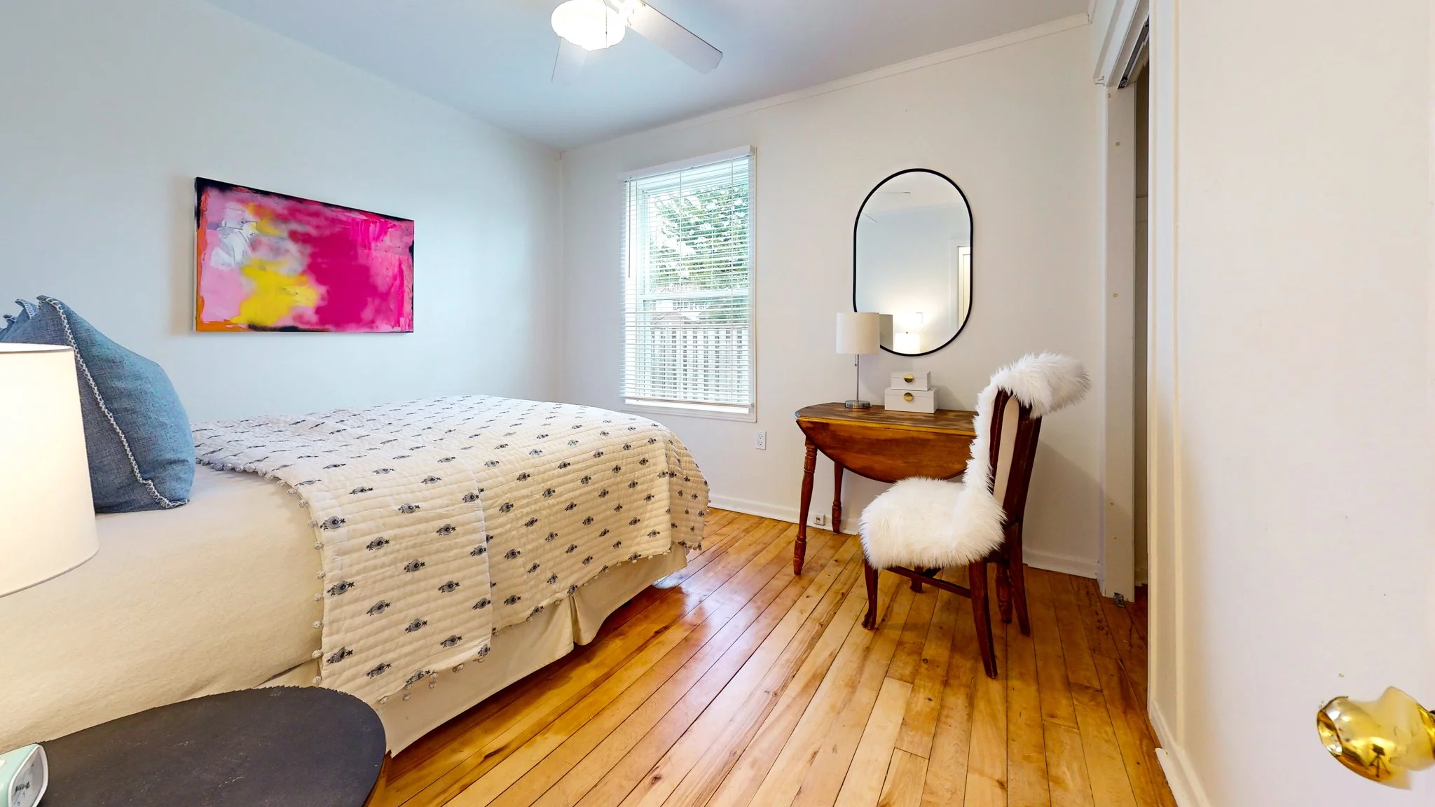 A bedroom with a bed, a wooden desk with a mirror and a fur-covered chair, a window with blinds, and a wall-mounted colorful abstract painting.