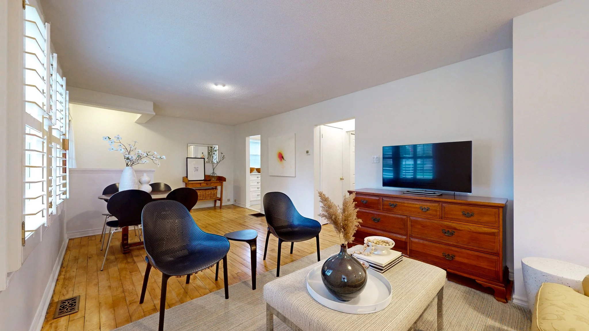 Living room with wooden floors, black chairs, a wooden dresser with a TV on top, a coffee table with decorative plants, and a dining area with a round table and black chairs.