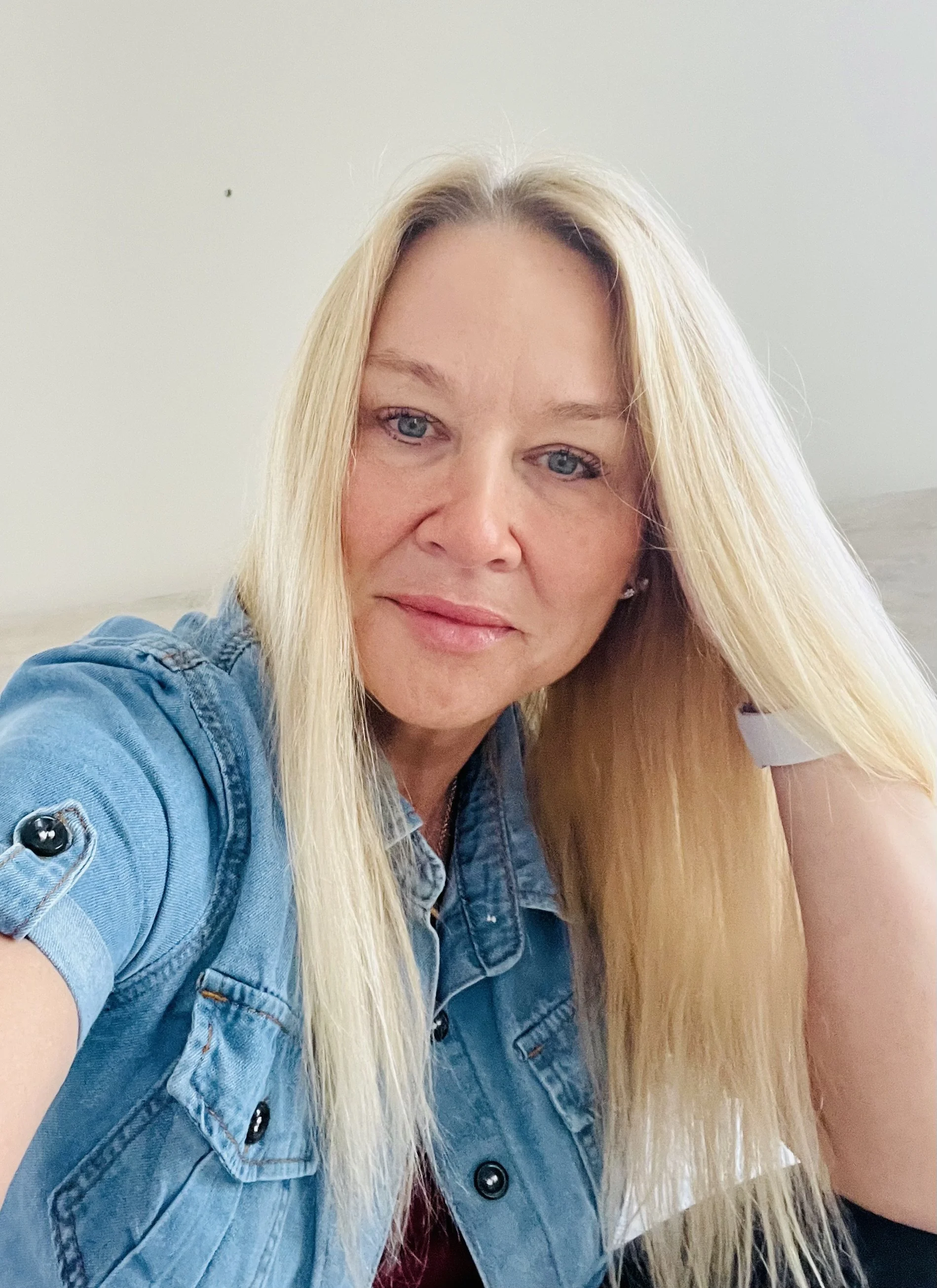 A woman with long blonde hair wearing a denim jacket, smiling softly at the camera.