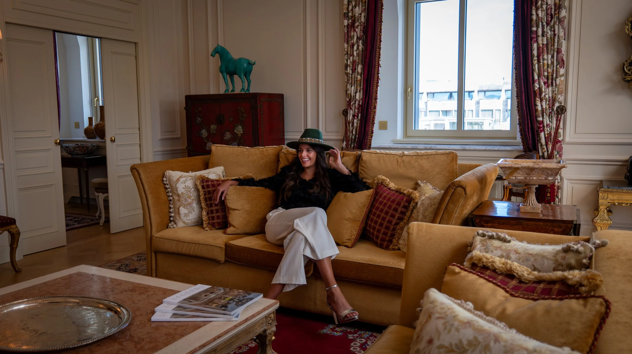 A woman sitting on a yellow sofa in a luxurious living room with classic decor, wearing a black top, white pants, a green hat, and smiling.