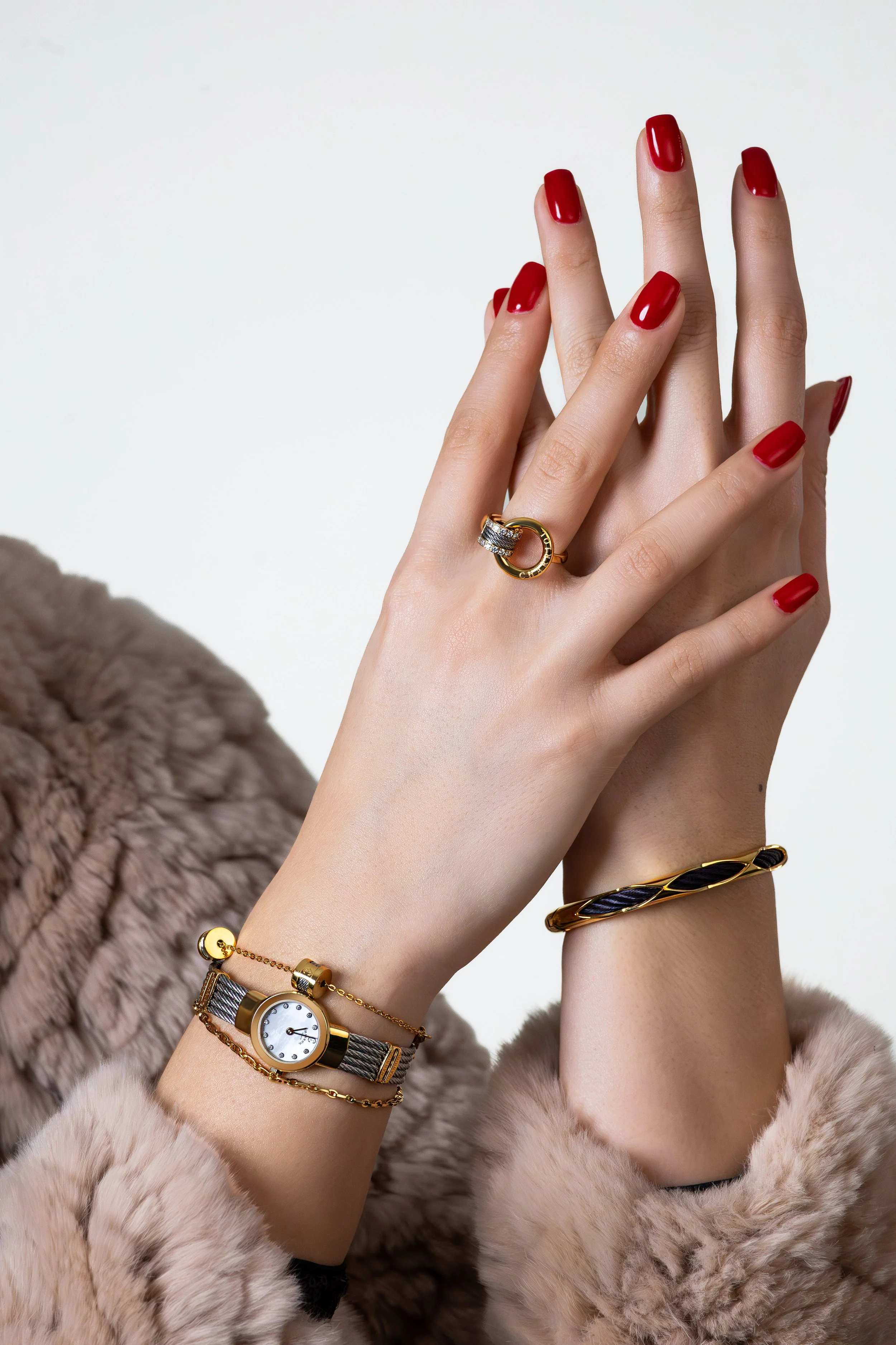 A person's hands with red painted nails and jewelry, including a watch, ring, bracelet, and cuff bracelet, against a plain background and wearing a fur coat.