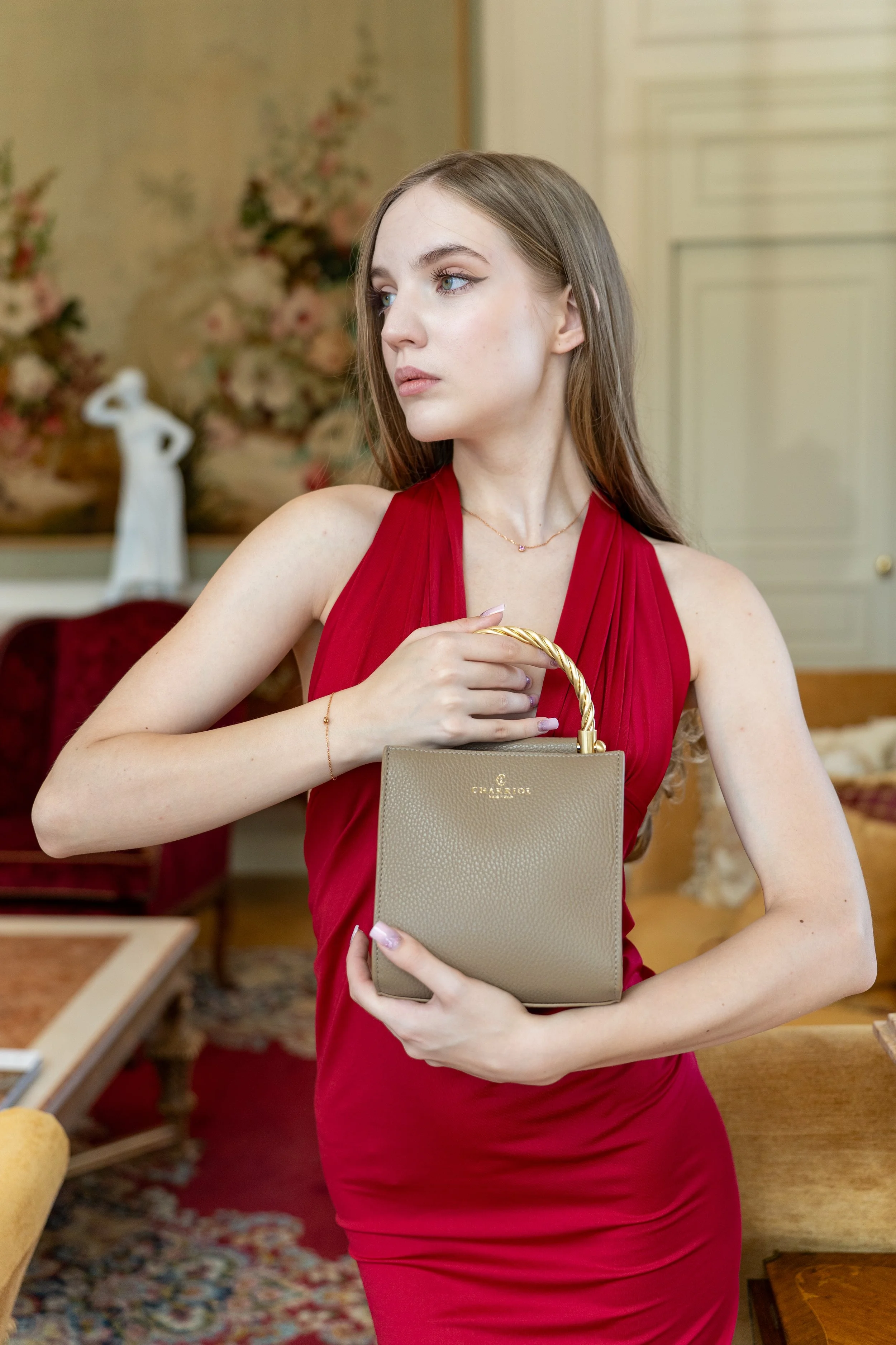 A woman in a red dress holding a beige handbag with a gold handle in an elegantly decorated room.