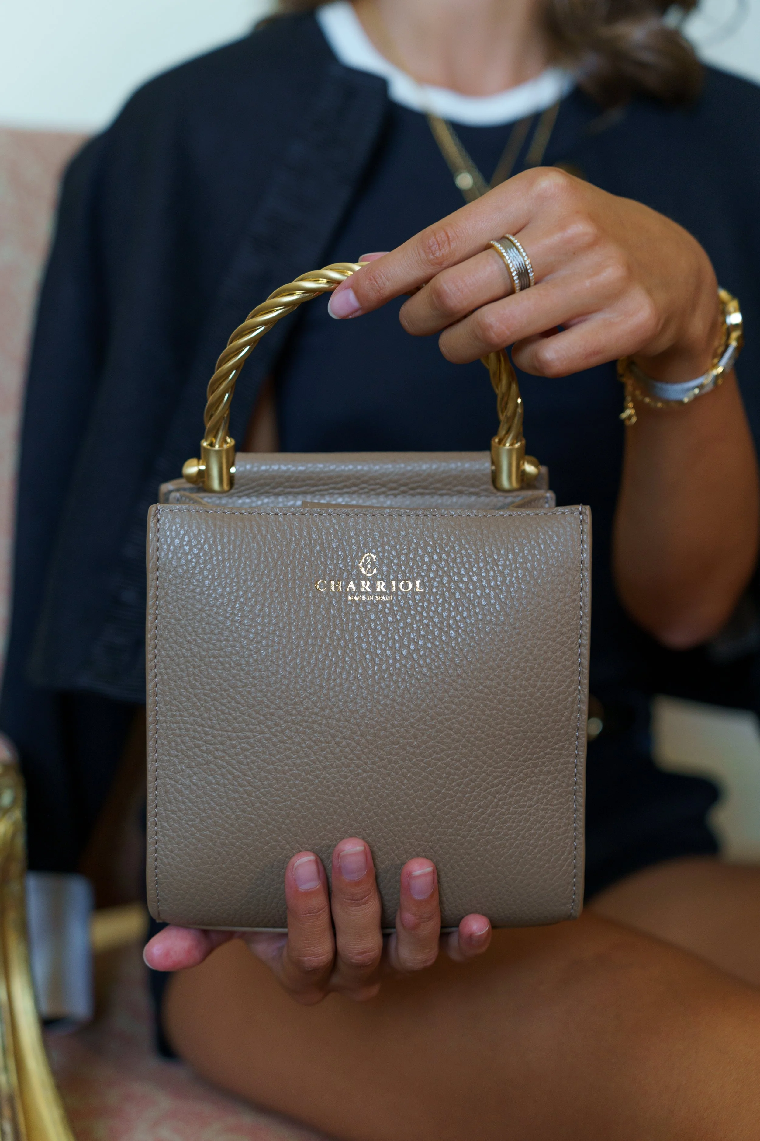 Person holding a beige purse with a gold twisted handle. The purse has a gold logo "CHARIOL" on the front. The person is wearing jewelry, including rings and bracelets.