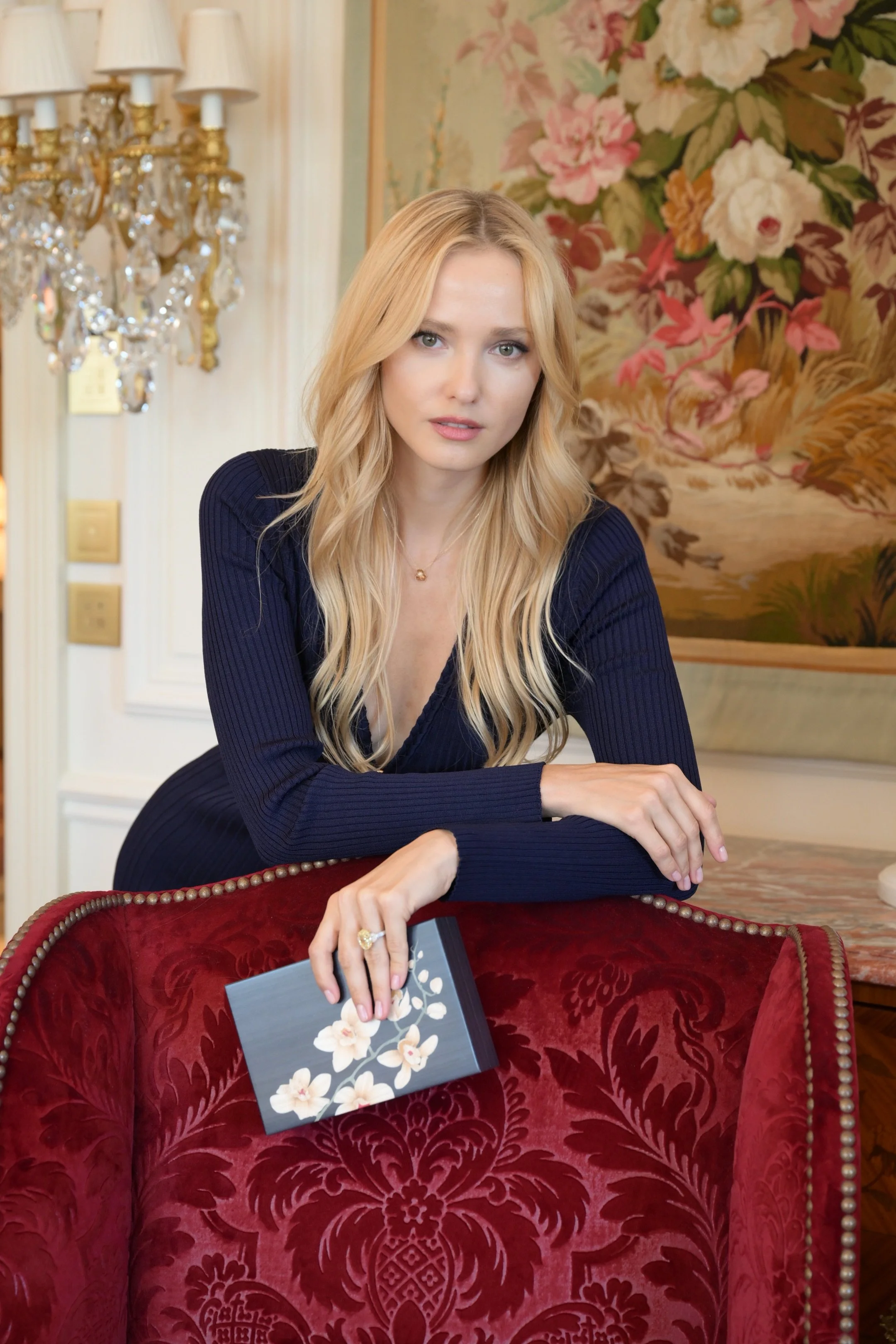 A blonde woman in a navy blue dress leaning on a red velvet armchair, holding a dark blue book with white flowers on the cover, in a decorated room with a floral wallpaper, a chandelier, and framed artwork.
