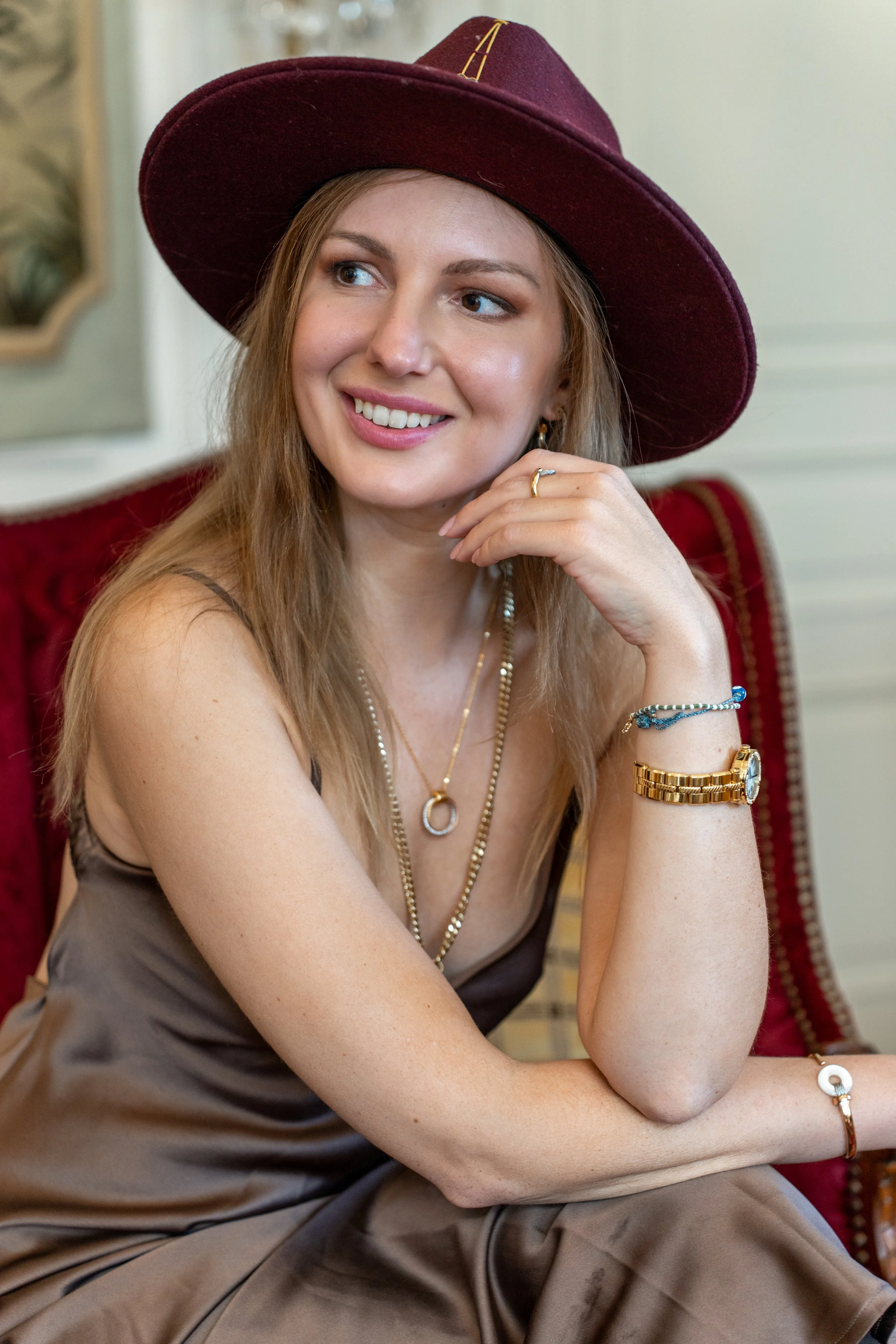 A young woman with light brown hair, wearing a large maroon hat and a satin brown dress, sitting on a red and gold upholstered chair, smiling and looking to the side, accessorized with jewelry including necklaces, rings, and a watch.