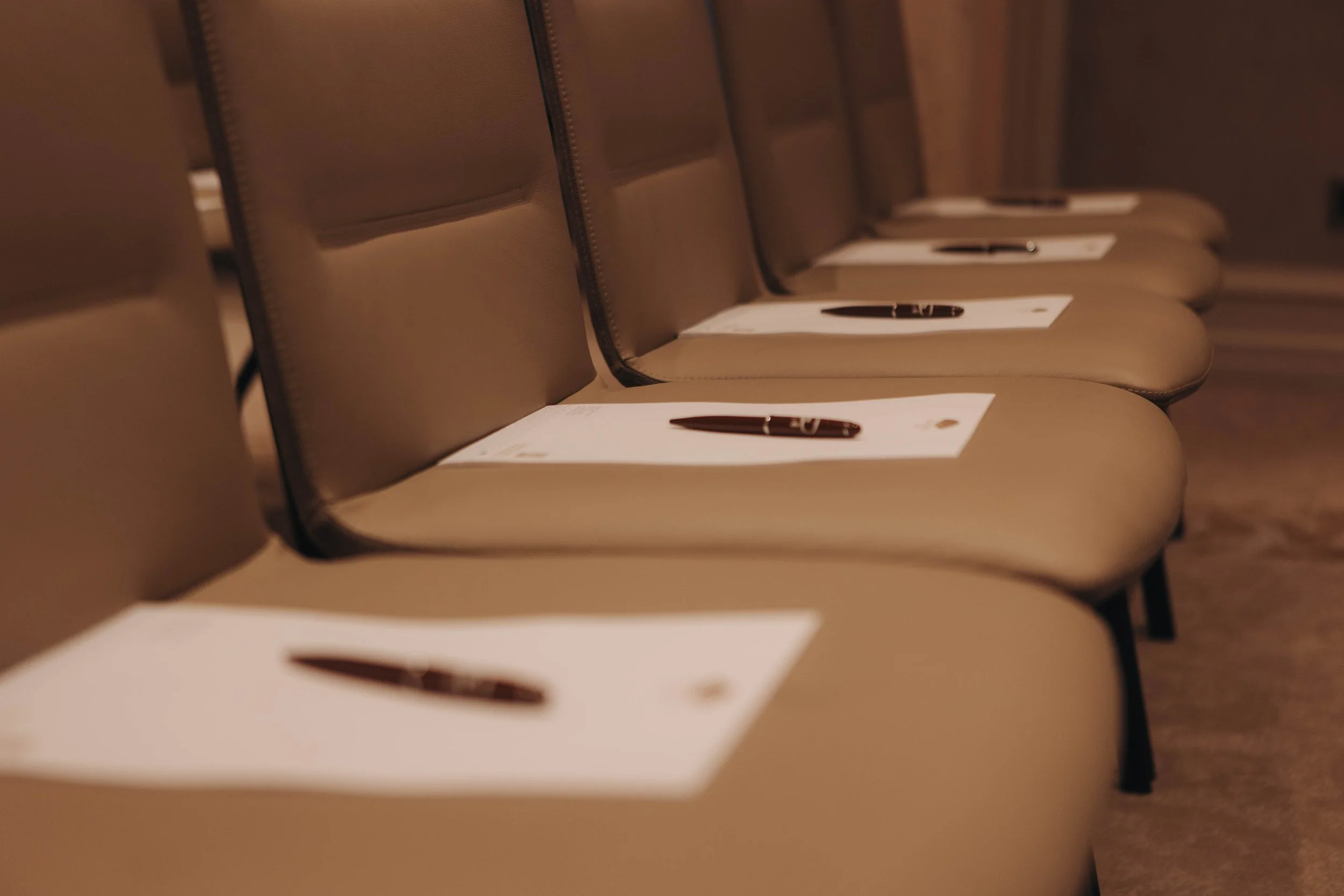 Multiple beige conference chairs arranged in a row, each with a pen and paper on the seat.
