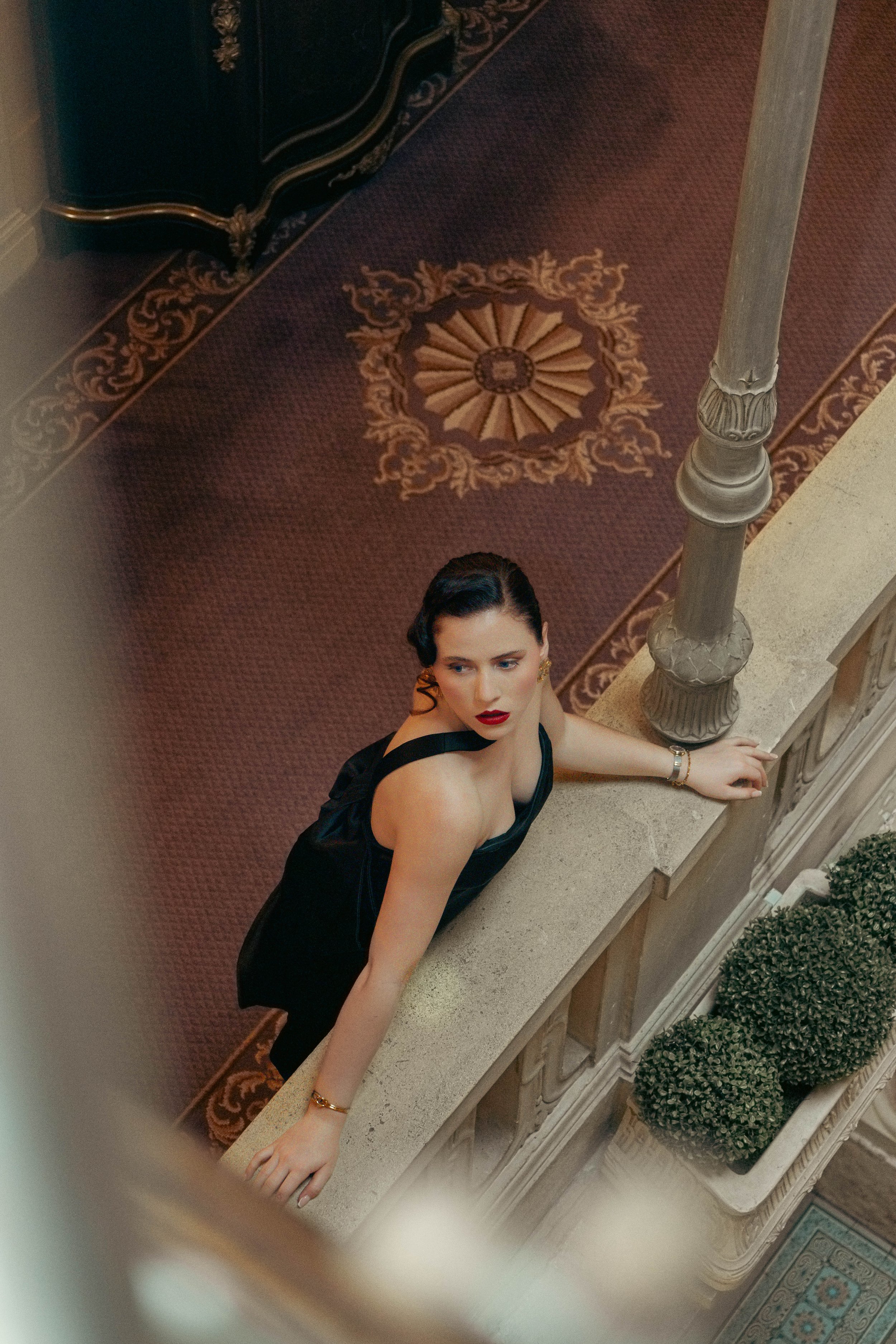 A woman with dark wavy hair, red lipstick, and wearing a black dress is looking upward while leaning on a stone balcony railing inside a luxurious building. The ornate carpet and decorative elements are visible in the background.