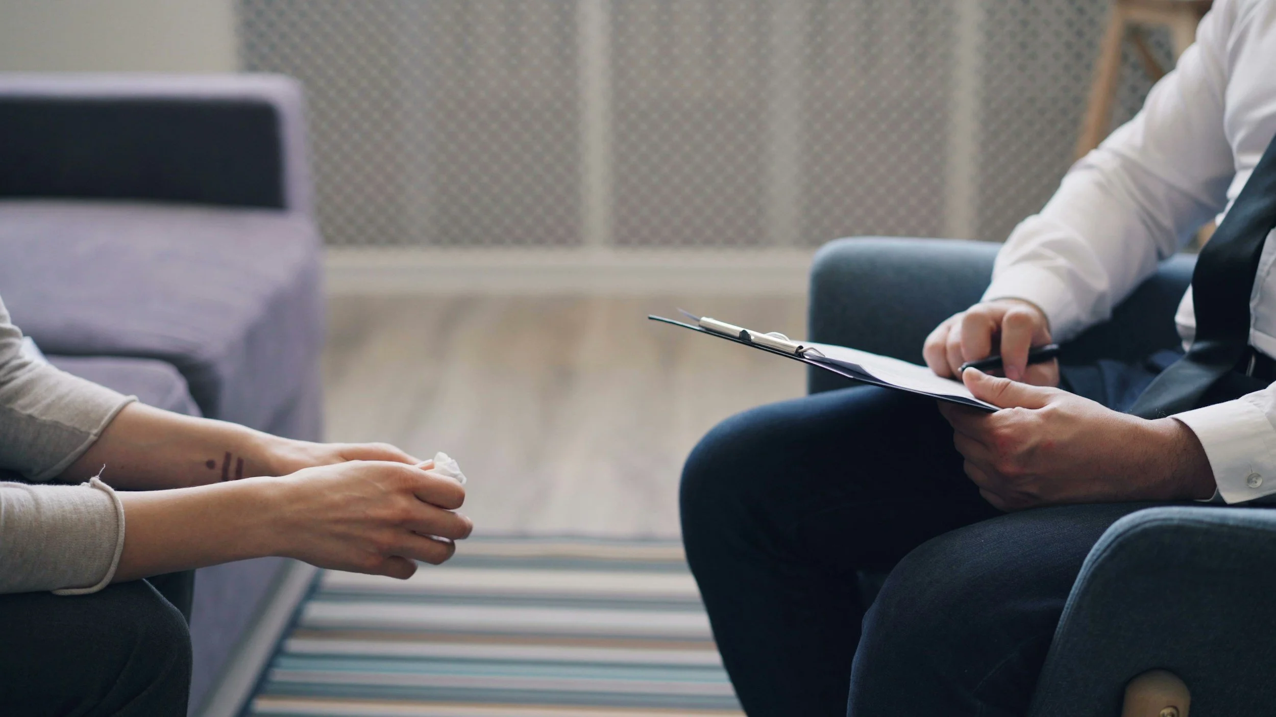 A person being interviewed by a counselor or therapist in a therapy session, with the person's hands resting on their lap and the counselor holding a clipboard and pen.