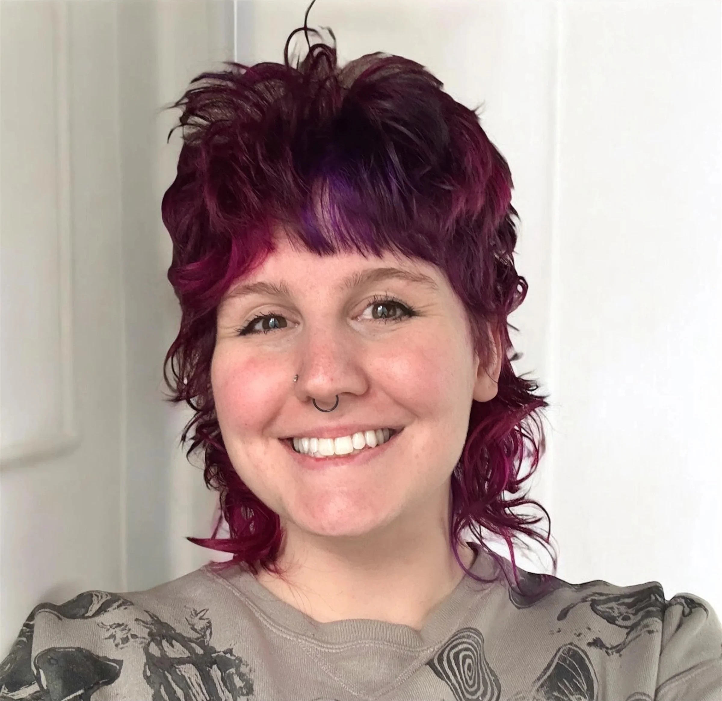 A woman with short, curly hair dyed in shades of purple and pink, smiling, with a nose piercing and septum ring, wearing a gray shirt with black abstract patterns, standing in front of a white door.