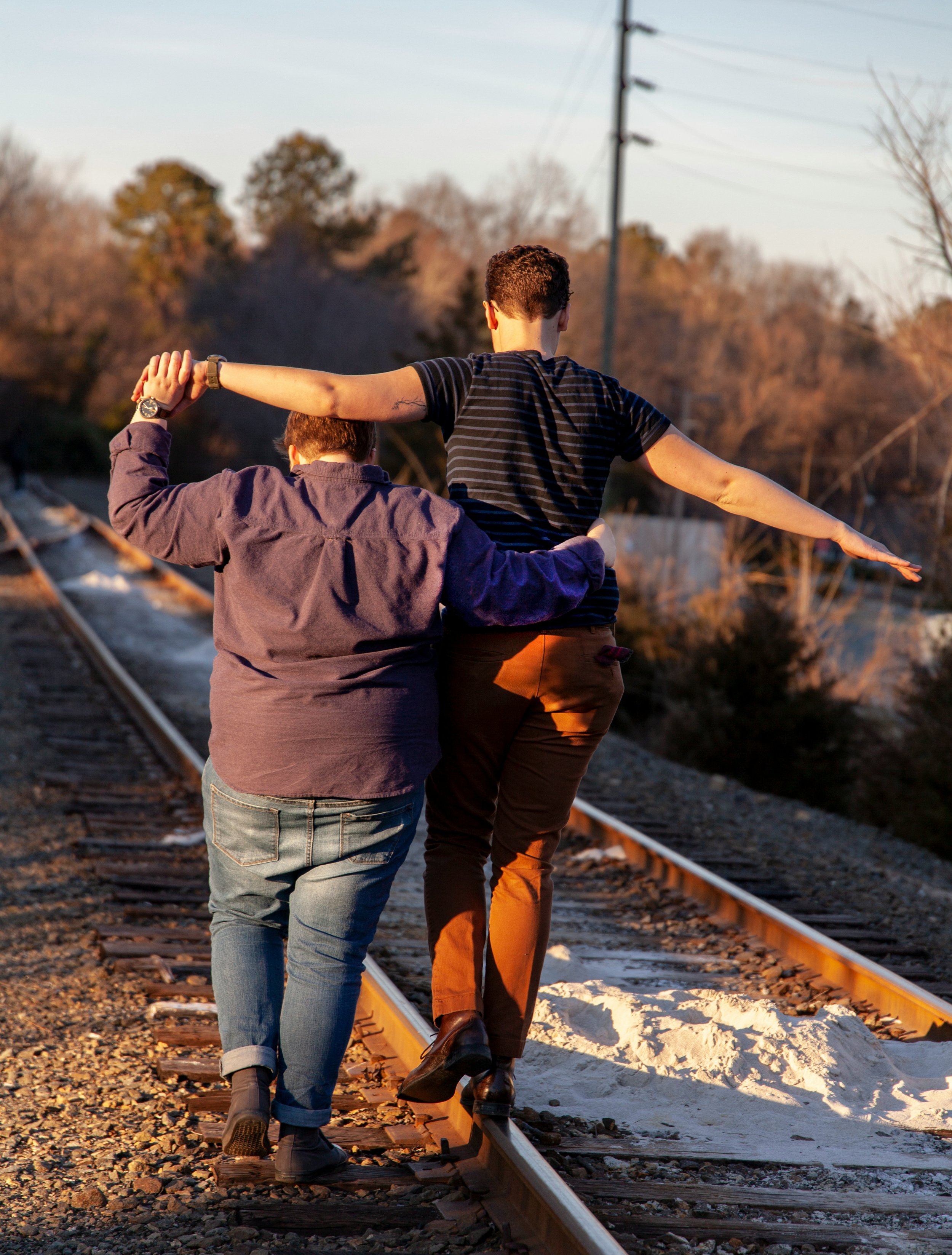 Two people walking on a train track with one person supporting the other, both seen from behind, during sunset with trees and power lines in the background.