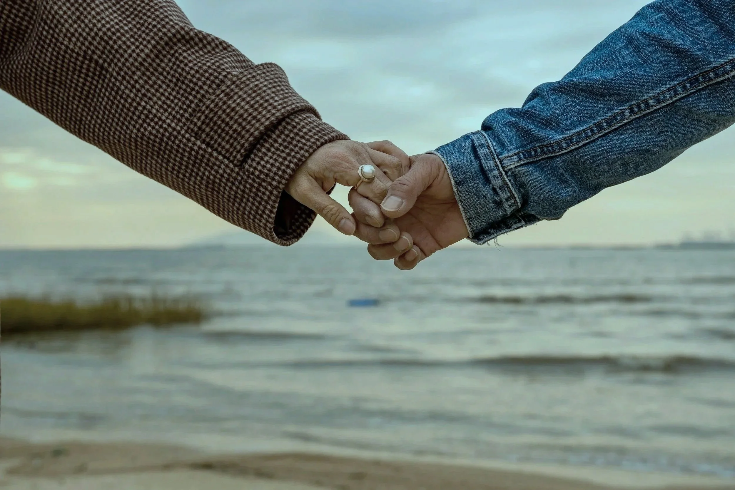Two people holding hands by the beach, one wearing a checkered jacket with a pearl ring and the other wearing a denim jacket.