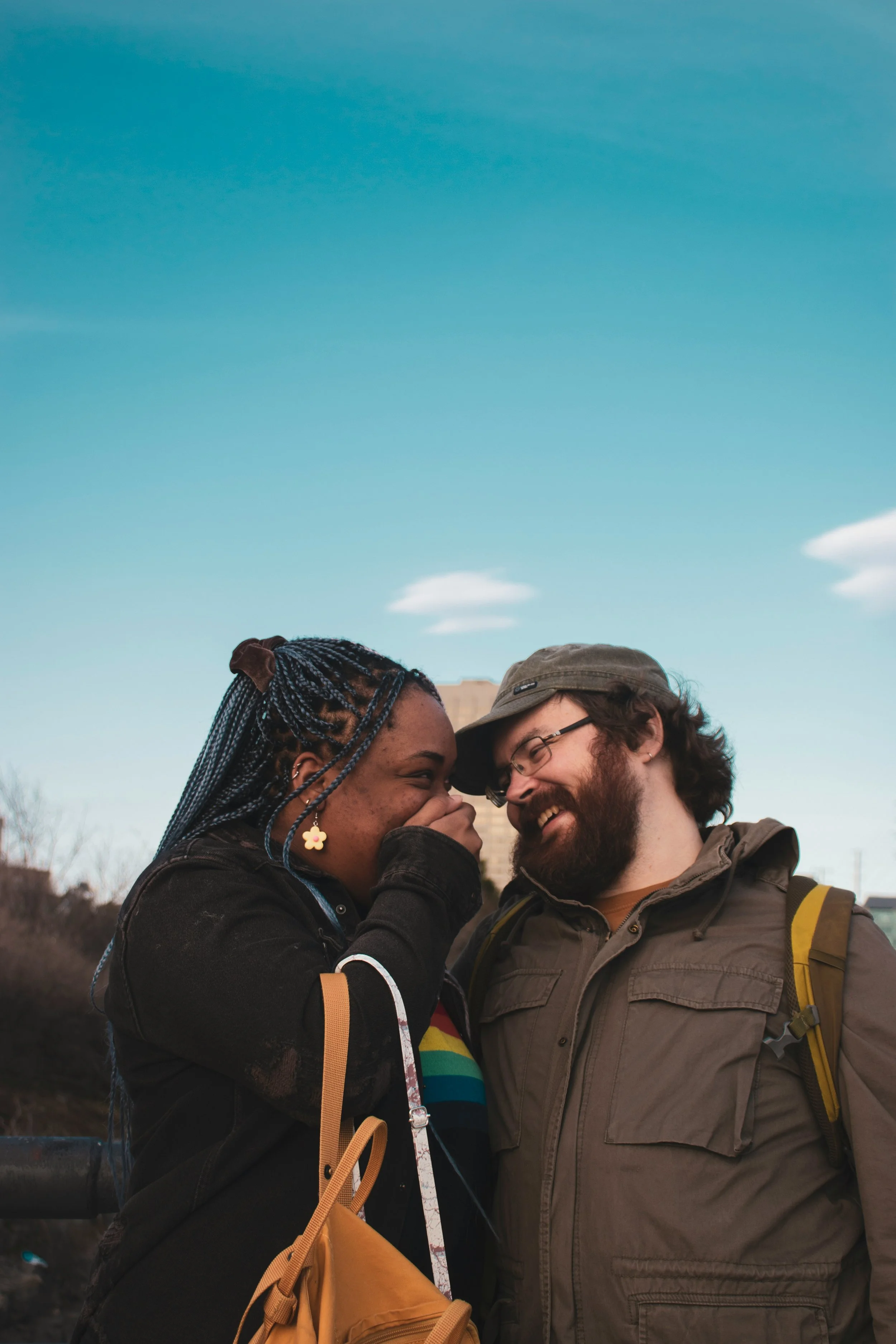 A man and a woman are smiling and whispering to each other outdoors against a blue sky with some clouds, sharing a joyful moment.