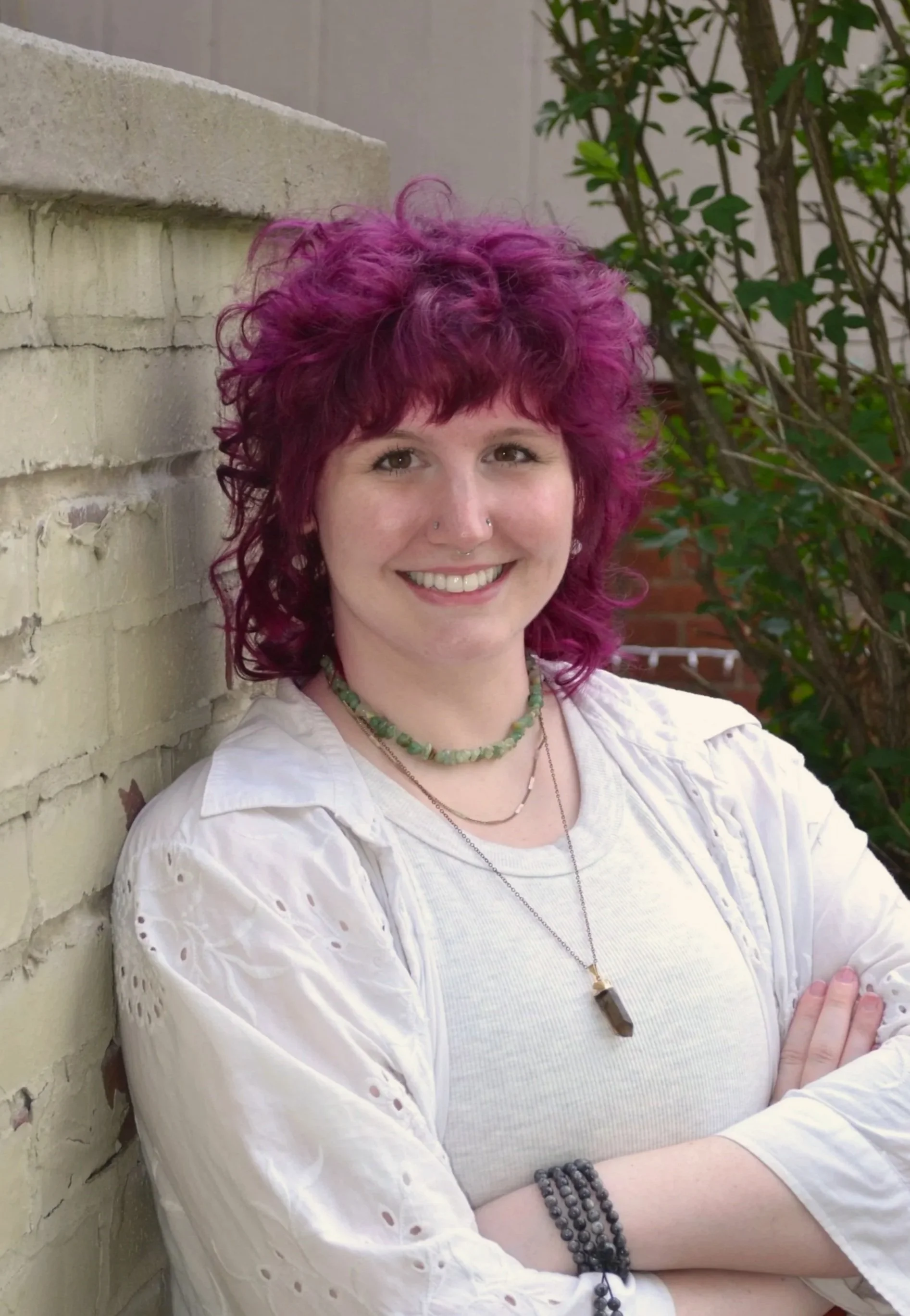 A person with curly purple hair smiling and standing against a brick wall with greenery in the background.