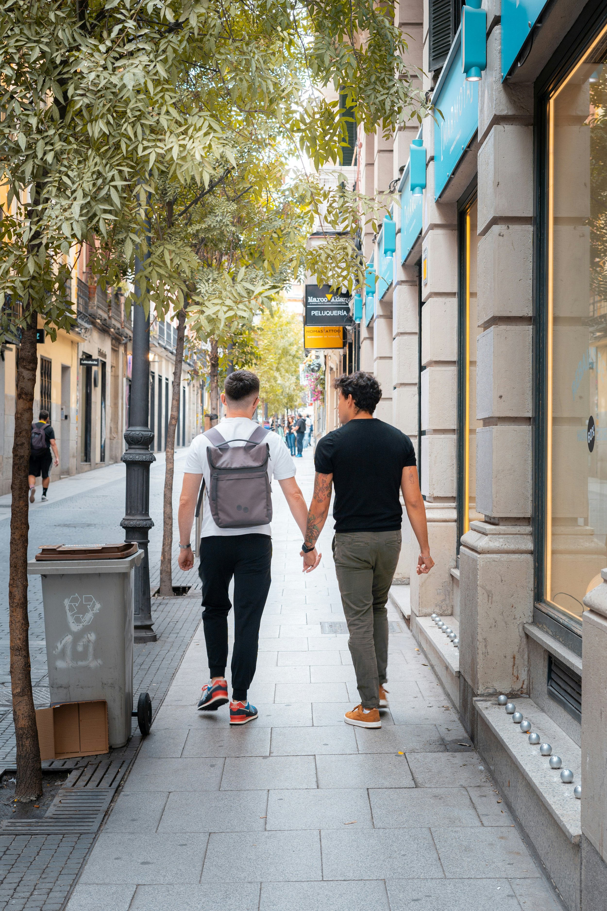 Two men walking down a paved city sidewalk, hand-in-hand, seen from behind, with trees on the left and storefronts on the right.
