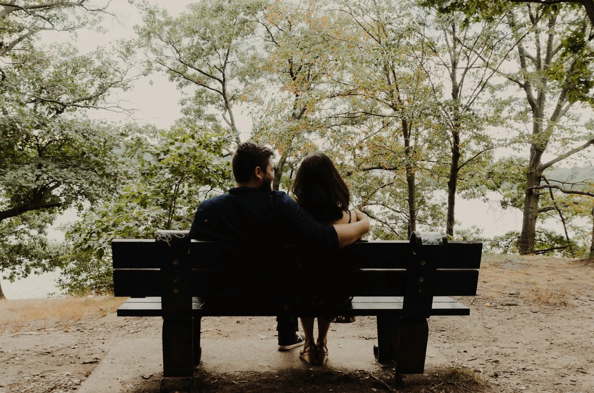 A couple sitting on a park bench embracing, with trees and a body of water in the background.