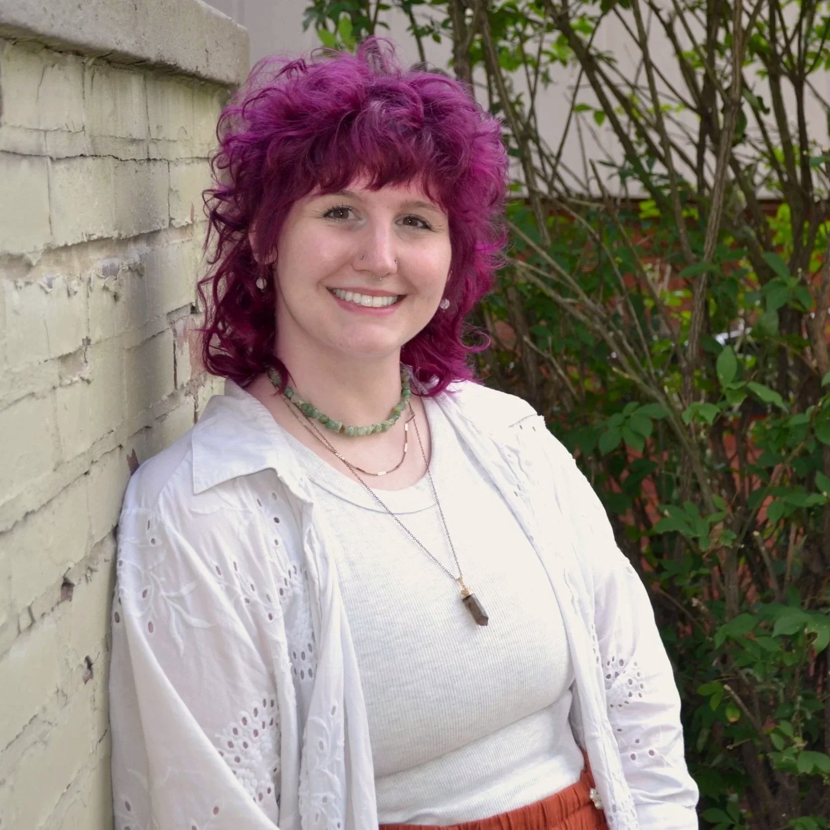 A person with curly purple hair leaning against a white brick wall outdoors, smiling, wearing a white eyelet blouse, layered necklaces, and a crystal pendant necklace, with green foliage in the background.