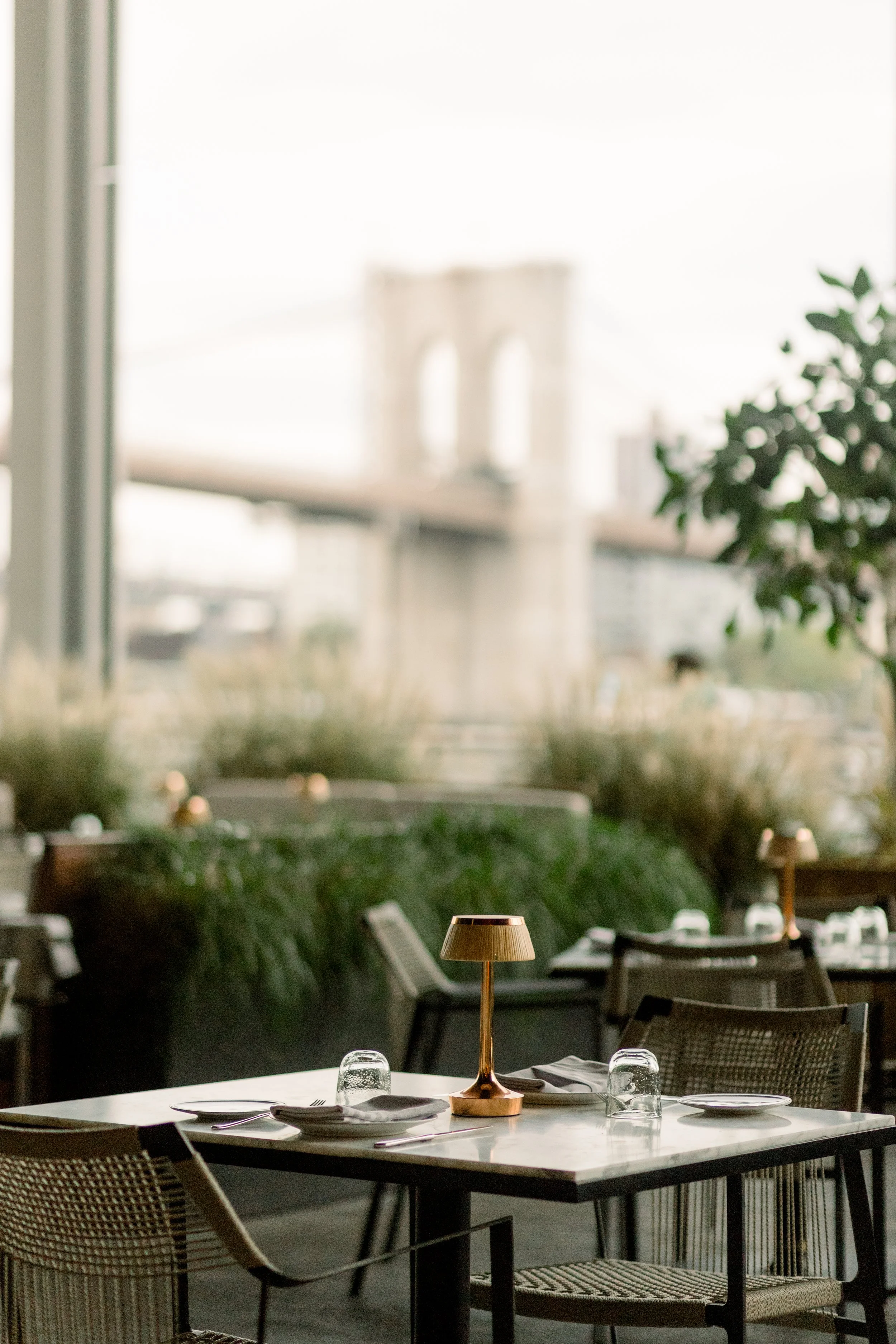 A restaurant interior with a table set for dining, including a small lamp, glasses, and utensils. Large windows in the background reveal blurred views of an outdoor bridge and greenery. The space has modern decor with chairs and a lush green plant.
