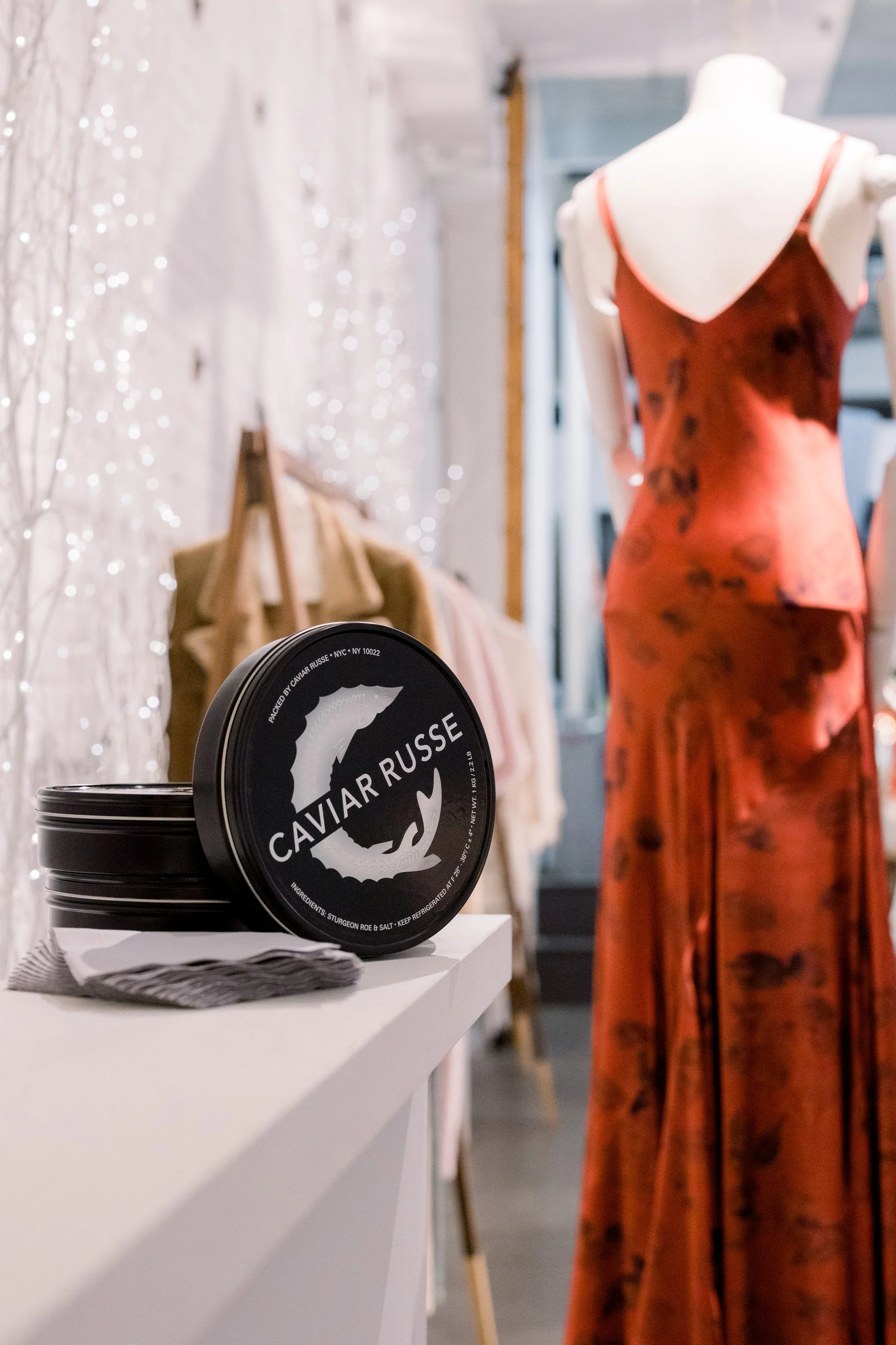 Close-up of a stack of black tins labeled 'Caviar Russe' on a white surface in a retail setting, with a blurred background of mannequins and a white wall decorated with fairy lights.