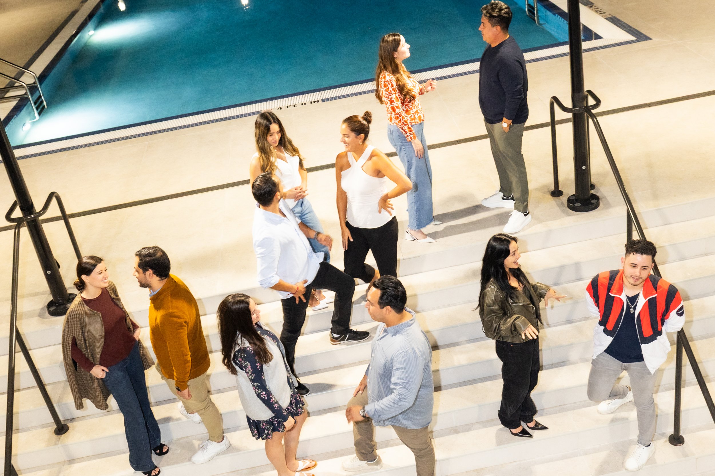 Group of people gathered on stairs near an indoor swimming pool, some engaged in conversation, others smiling and looking around.