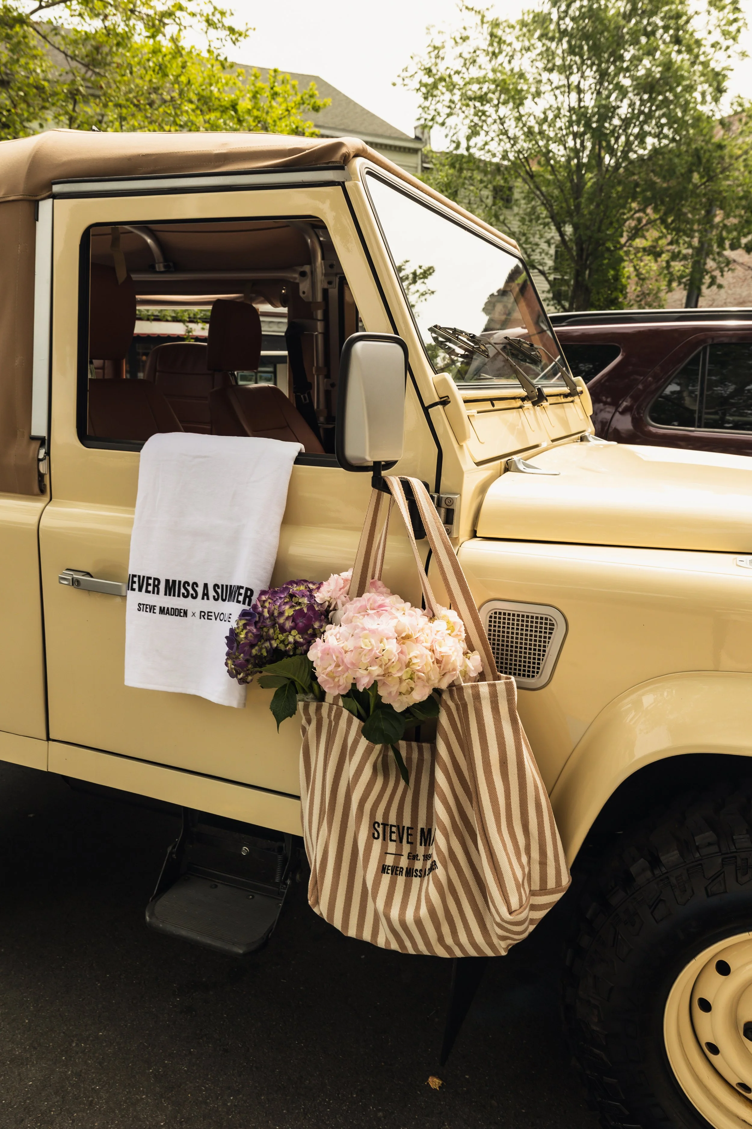 Yellow vintage off-road vehicle with a floral decoration and tote bags hanging on the side, parked outdoors with trees in the background.