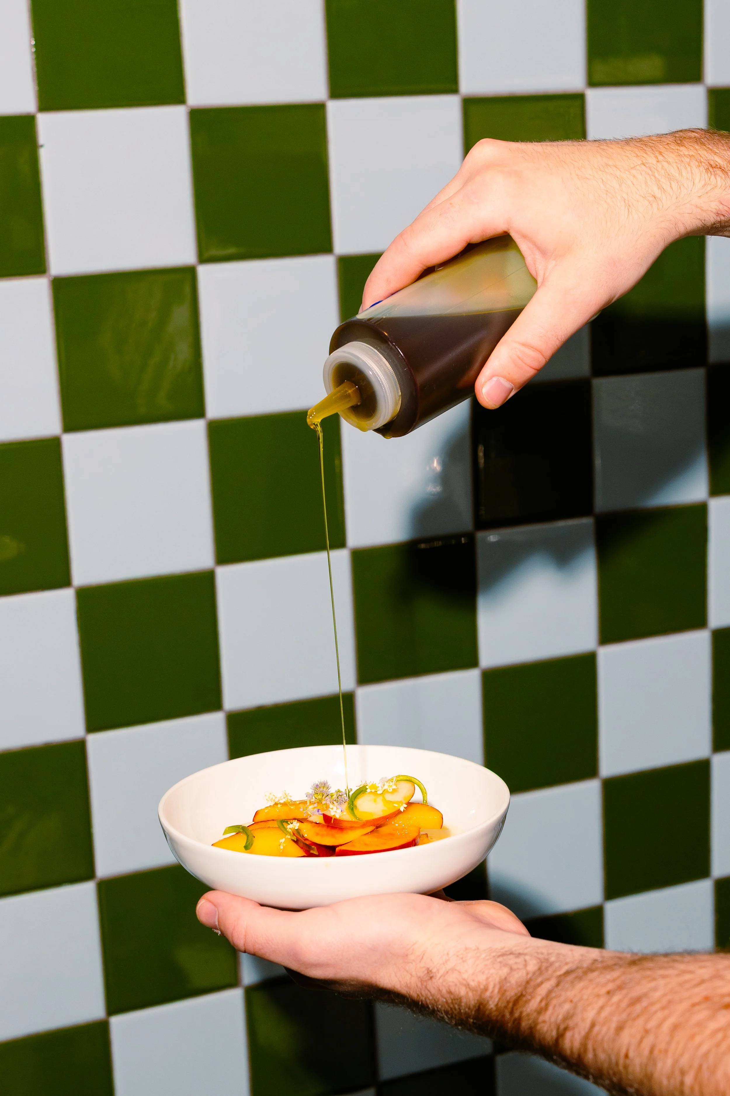 A hand holding a white bowl with a colorful salad, while another hand drizzles salad dressing from a squeeze bottle above the bowl. The background features a checkered green and white tiled wall.
