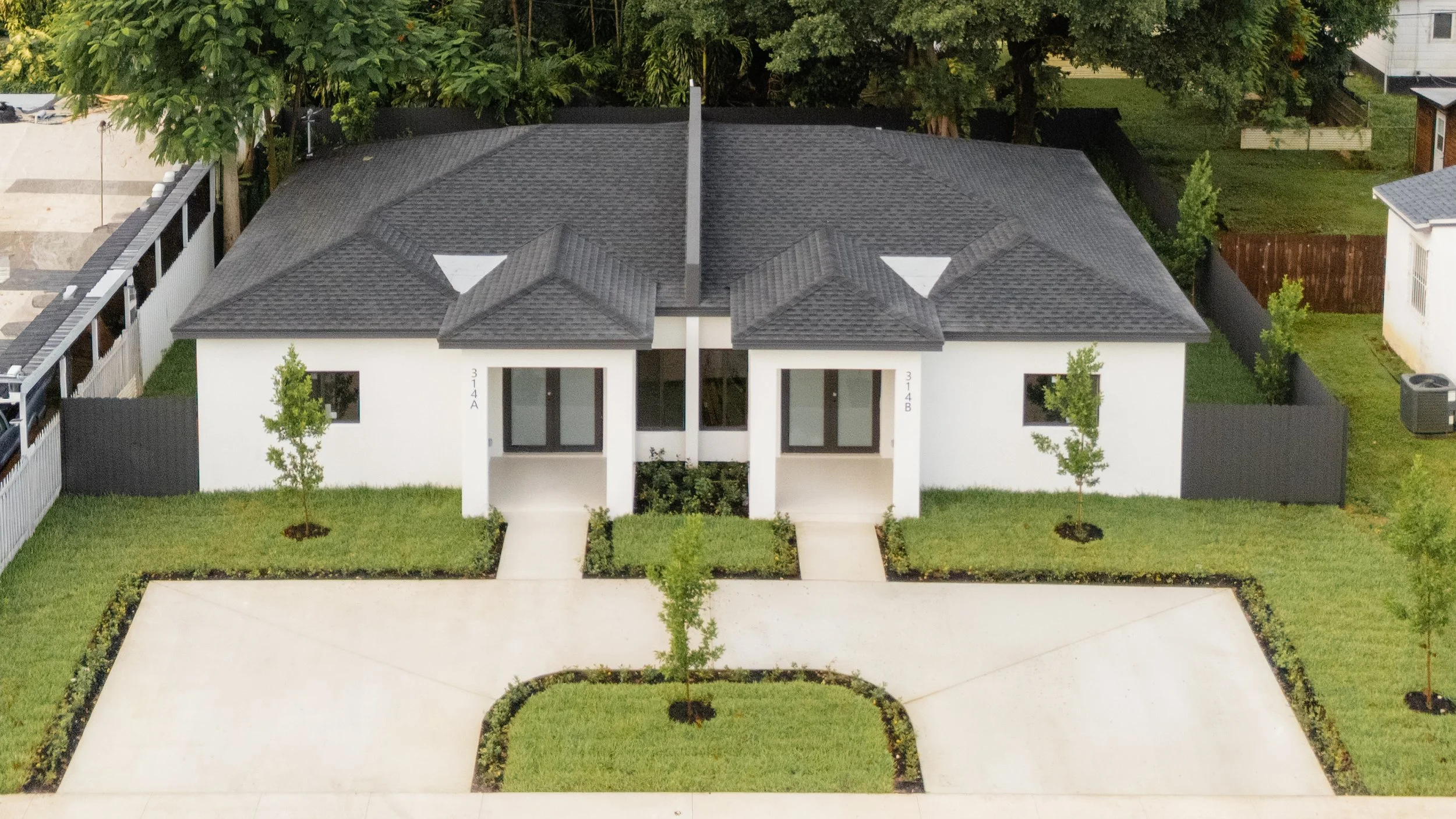 Aerial view of a modern duplex house with a black shingle roof, white exterior walls, small front porch, and a green lawn with young trees. The house has symmetrical design with two front doors, each with large glass panels. There are concrete pathways leading to the street, and neighboring houses and trees are visible around.