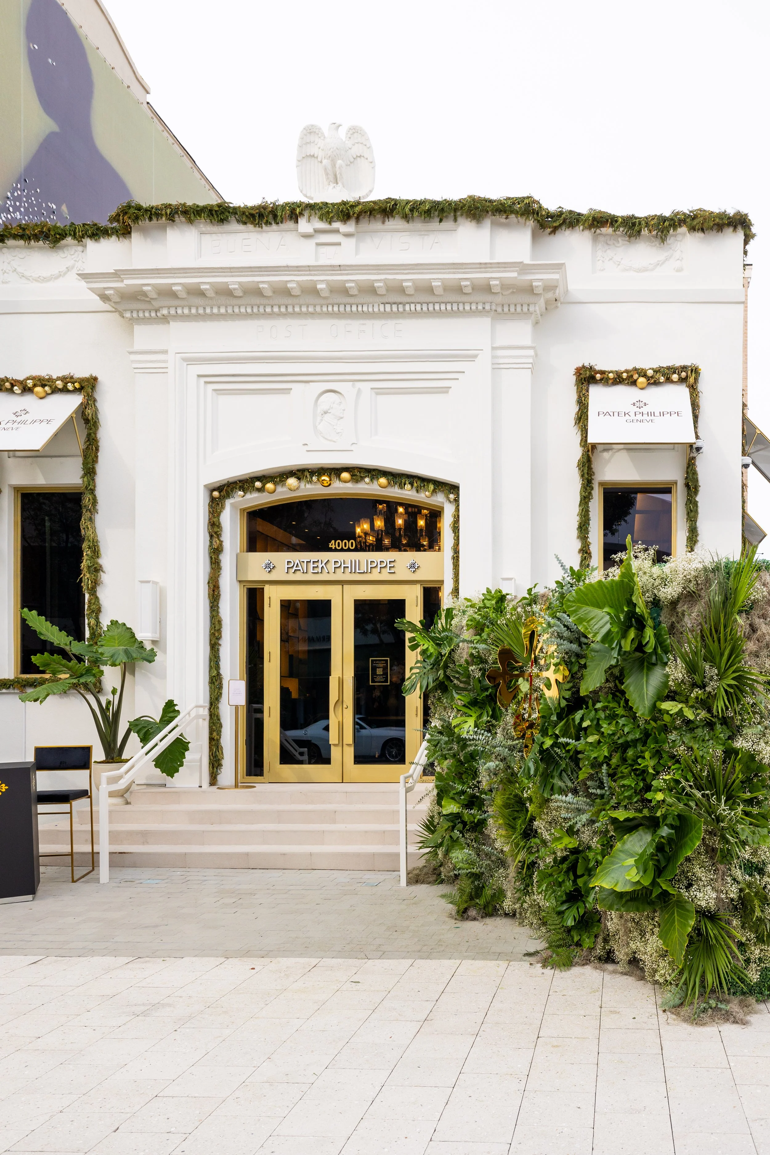 The exterior of a luxury watch store, Patek Philippe, decorated with holiday garlands and ornaments, with lush greenery and plants outside.