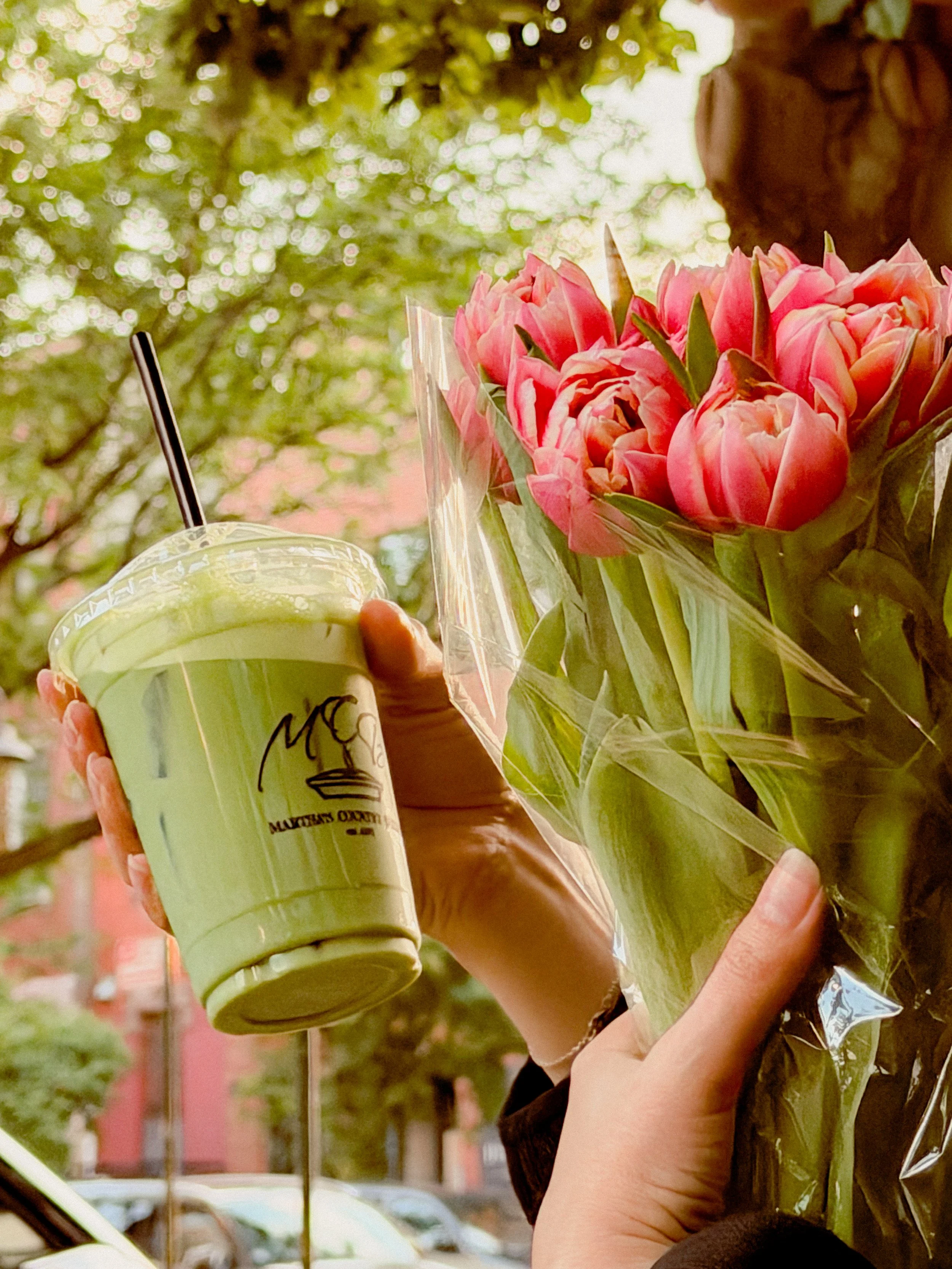 A person holding a bouquet of pink tulips wrapped in clear plastic. The person is holding a green cup with a straw in it, and the background shows trees and a building.