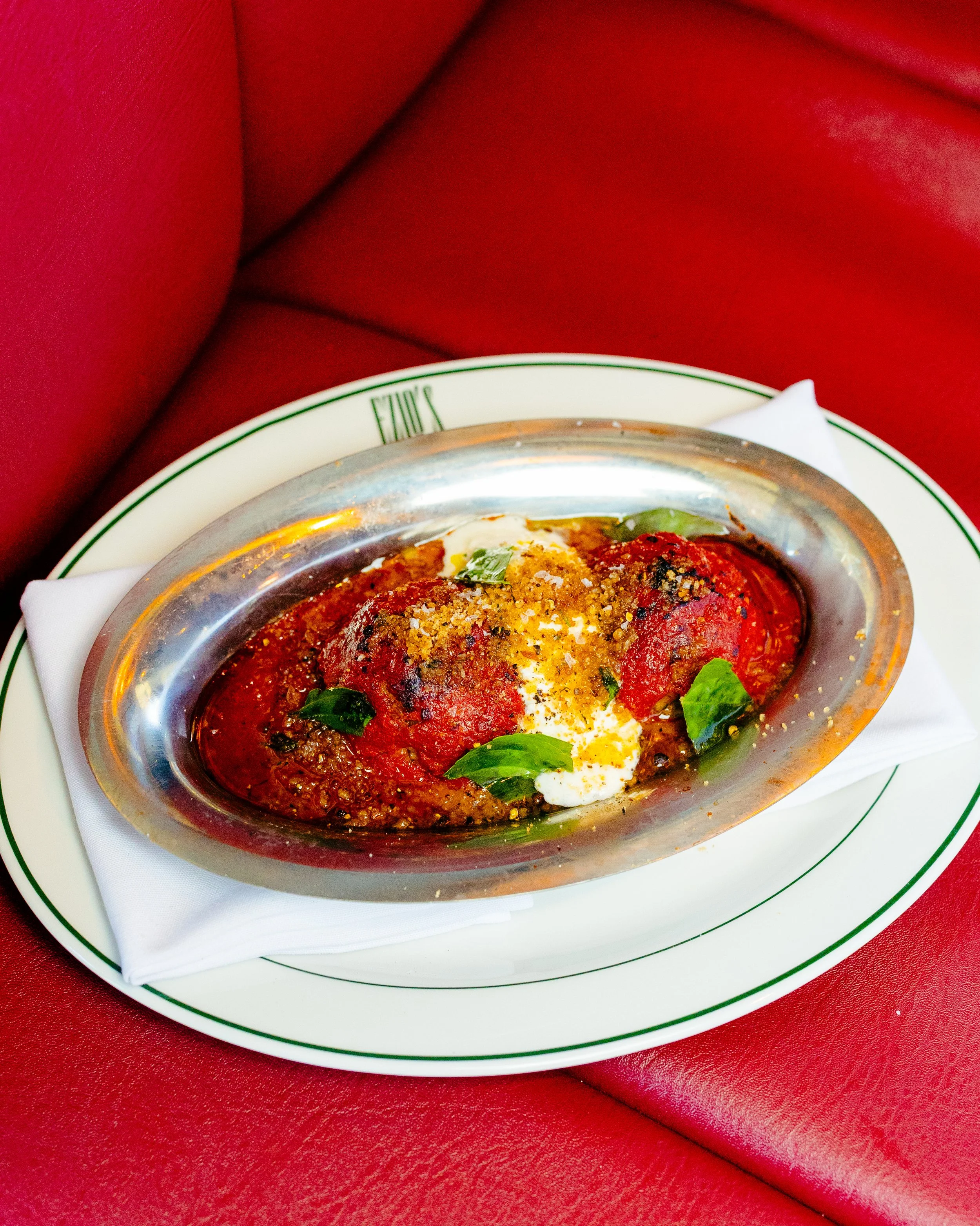 A plate of baked meat topped with tomato sauce, cheese, and herbs, served on a white plate with a green rim on a red table with red napkin.