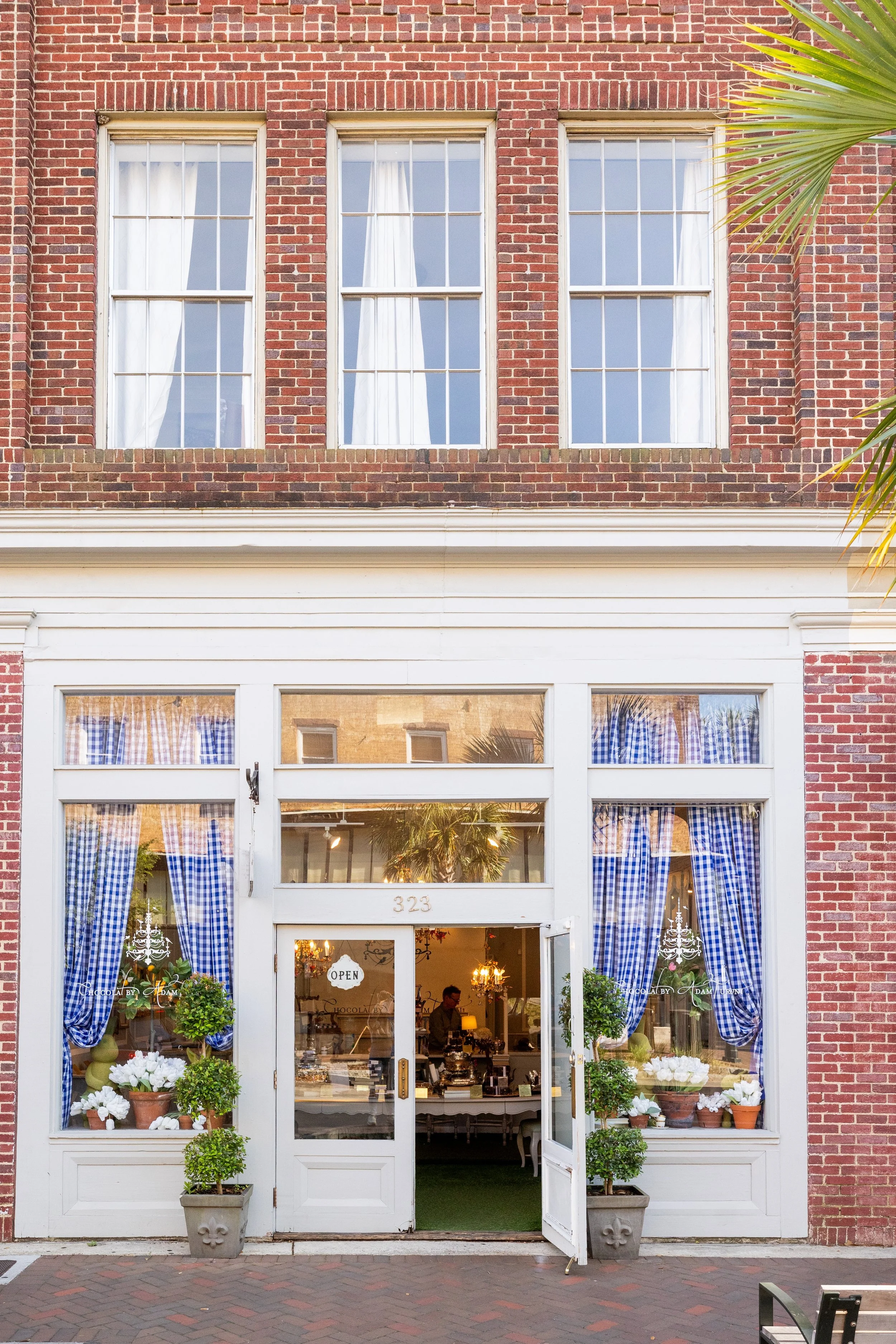 The storefront features a white wooden exterior with large glass windows covered by blue and white checkered curtains. It's decorated with potted plants and white flowers at the entrance. The door has an 'Open' sign, and the inside appears warmly lit with chandeliers and showcases items on display.