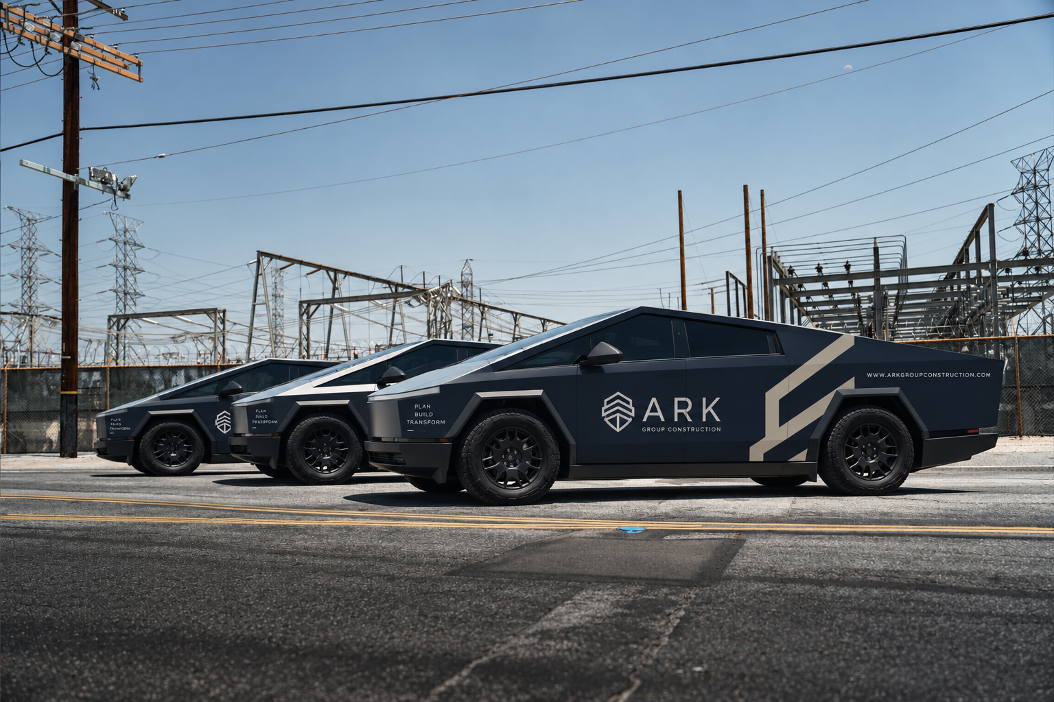Three futuristic black vehicles with angular designs parked on a city street, covered in branding for ARK Group Construction, with electrical towers and wires in the background under a clear blue sky.