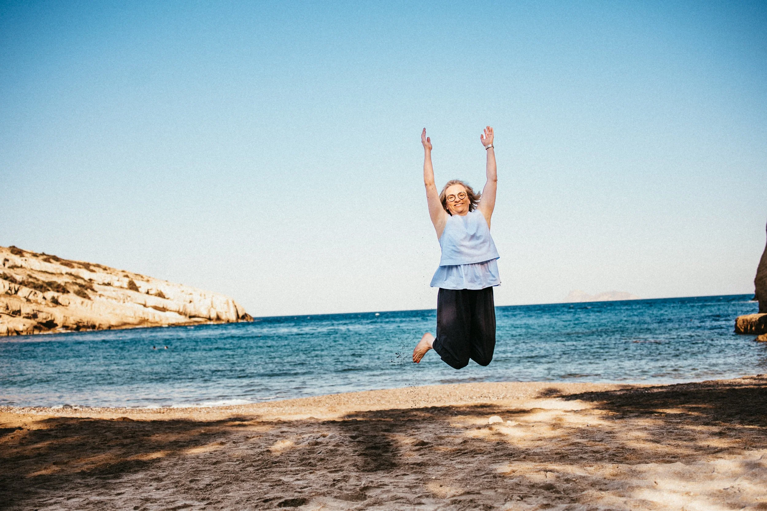 Frau springt freudig auf einem Strand mit Meer und Felsen im Hintergrund, sonniger Tag.