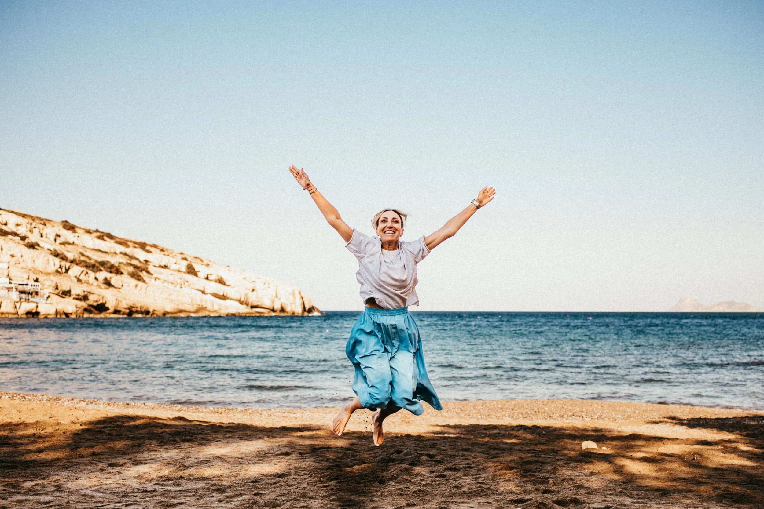 Eine fröhliche Frau springt barfuß am Strand bei Sonnenschein, im Hintergrund das Meer und eine Felsklippe.