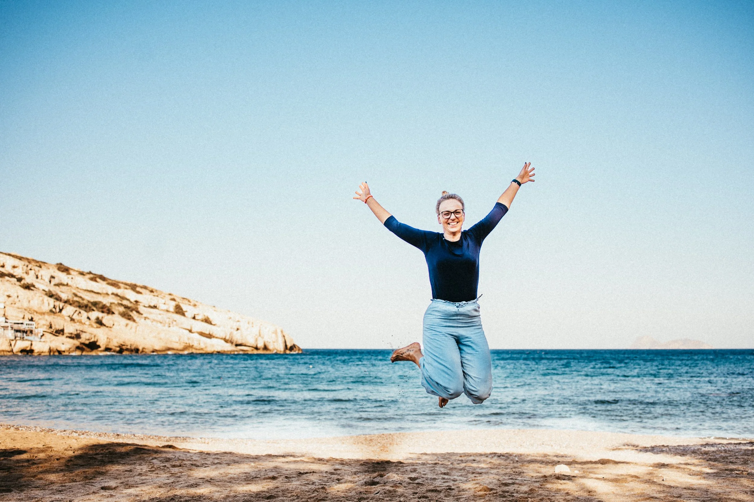 Frau springt glücklich am Strand mit Meer und Felsen im Hintergrund.